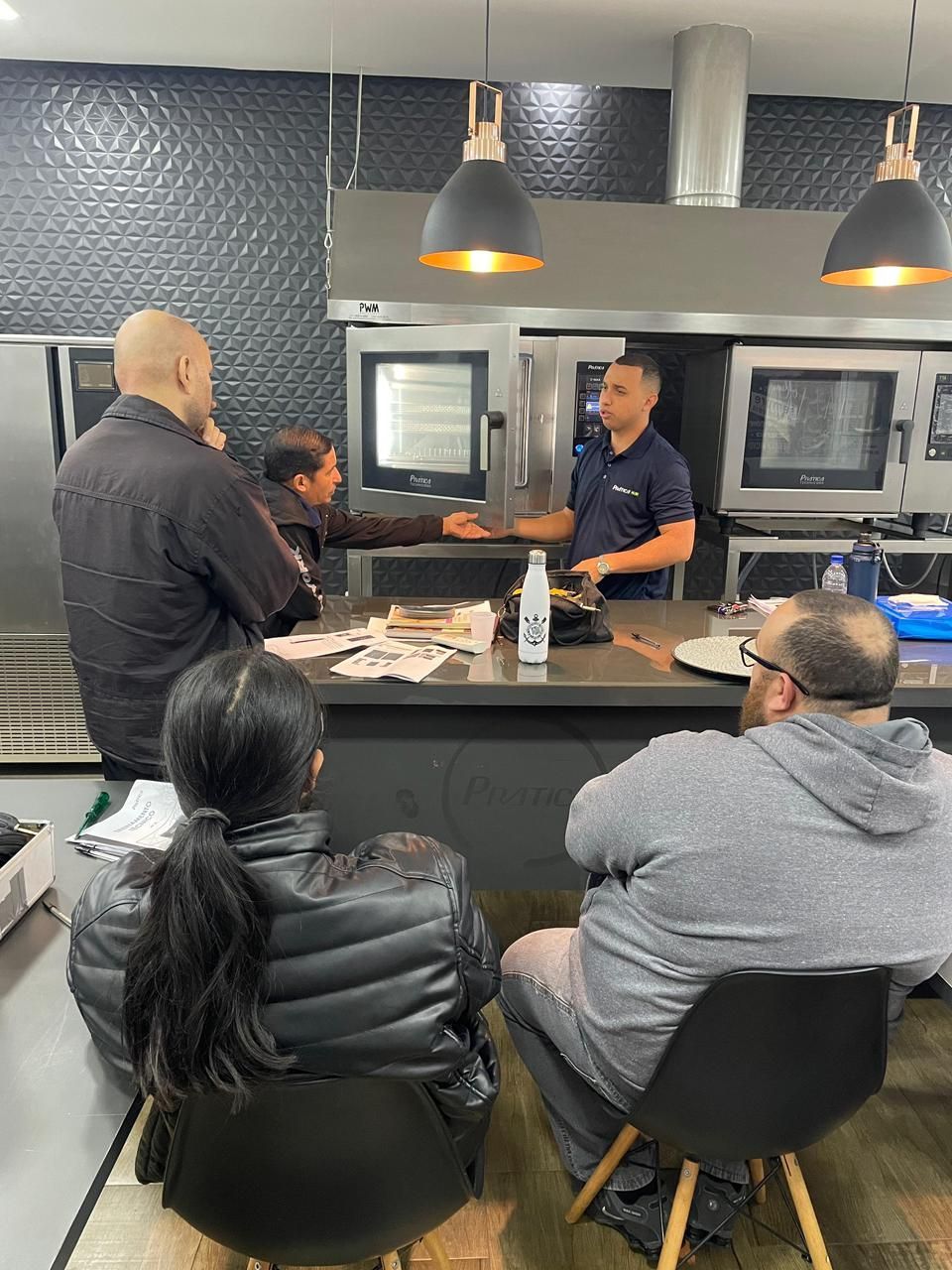 Um homem fala para um grupo em uma cozinha. Eles estão reunidos em volta de um balcão, perto de fornos.