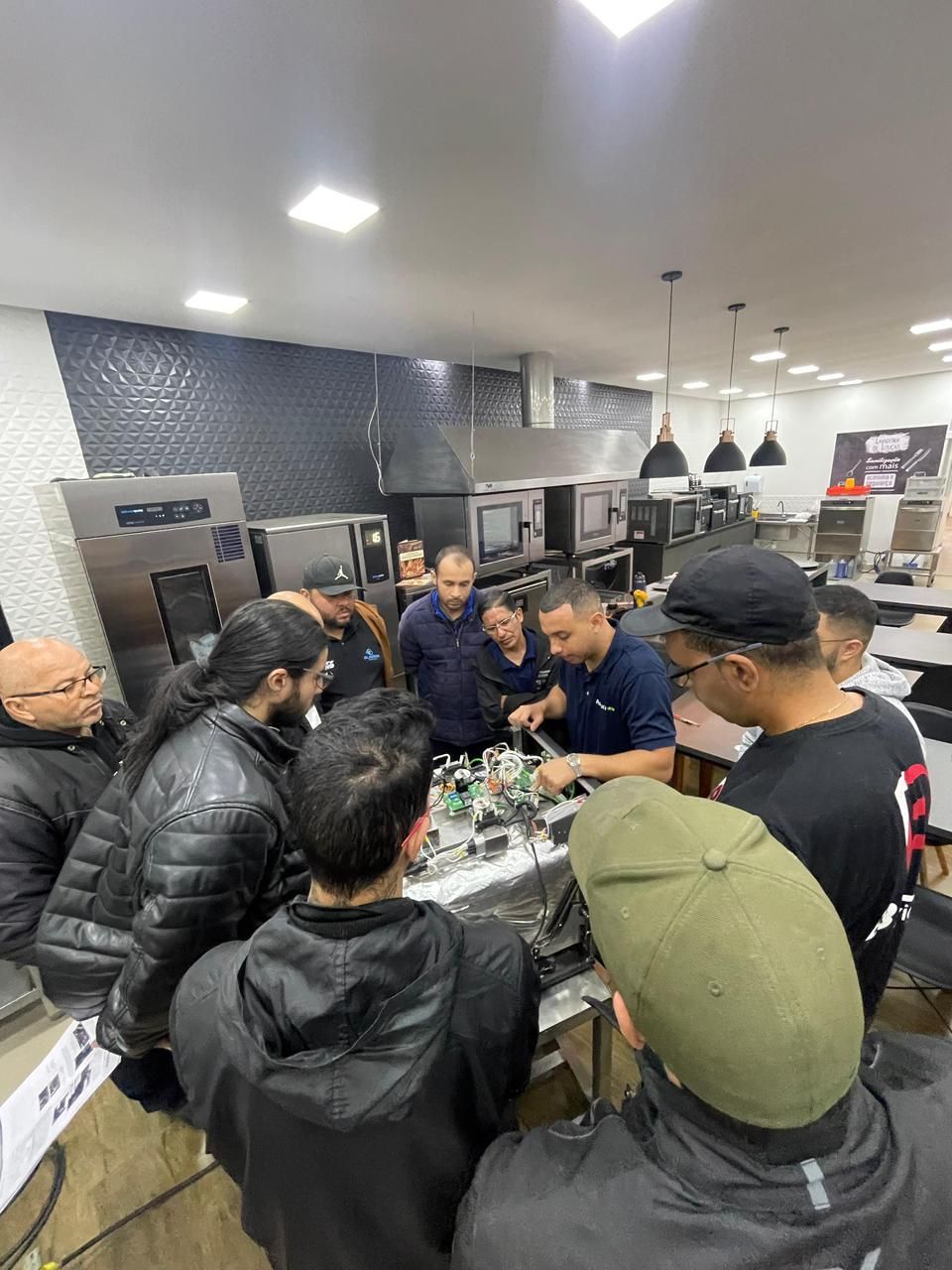 Grupo de pessoas em uma cozinha, observando um técnico trabalhando em um equipamento.
