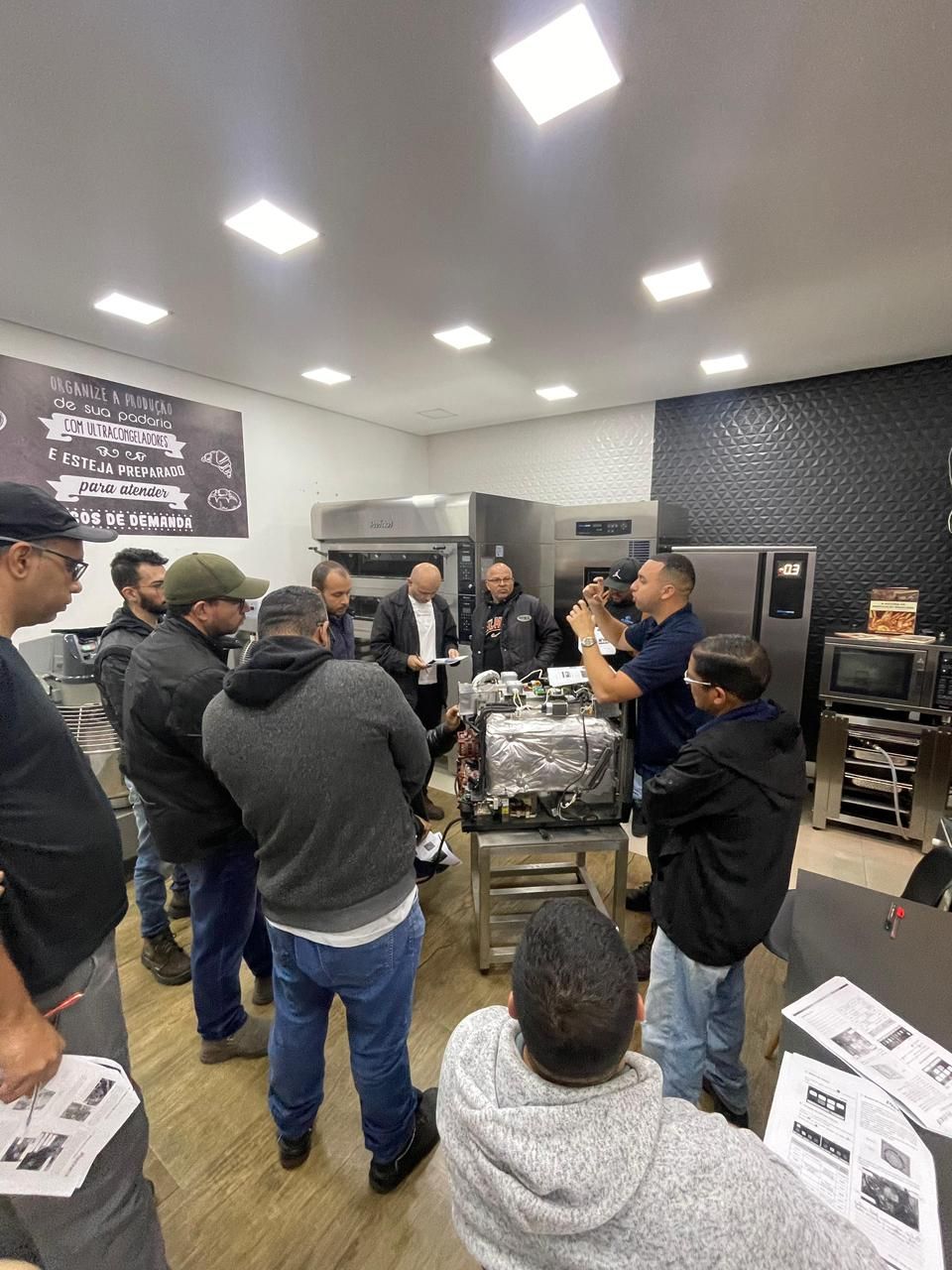 Grupo de pessoas observando uma demonstração, possivelmente uma oficina em uma cozinha comercial com equipamentos.