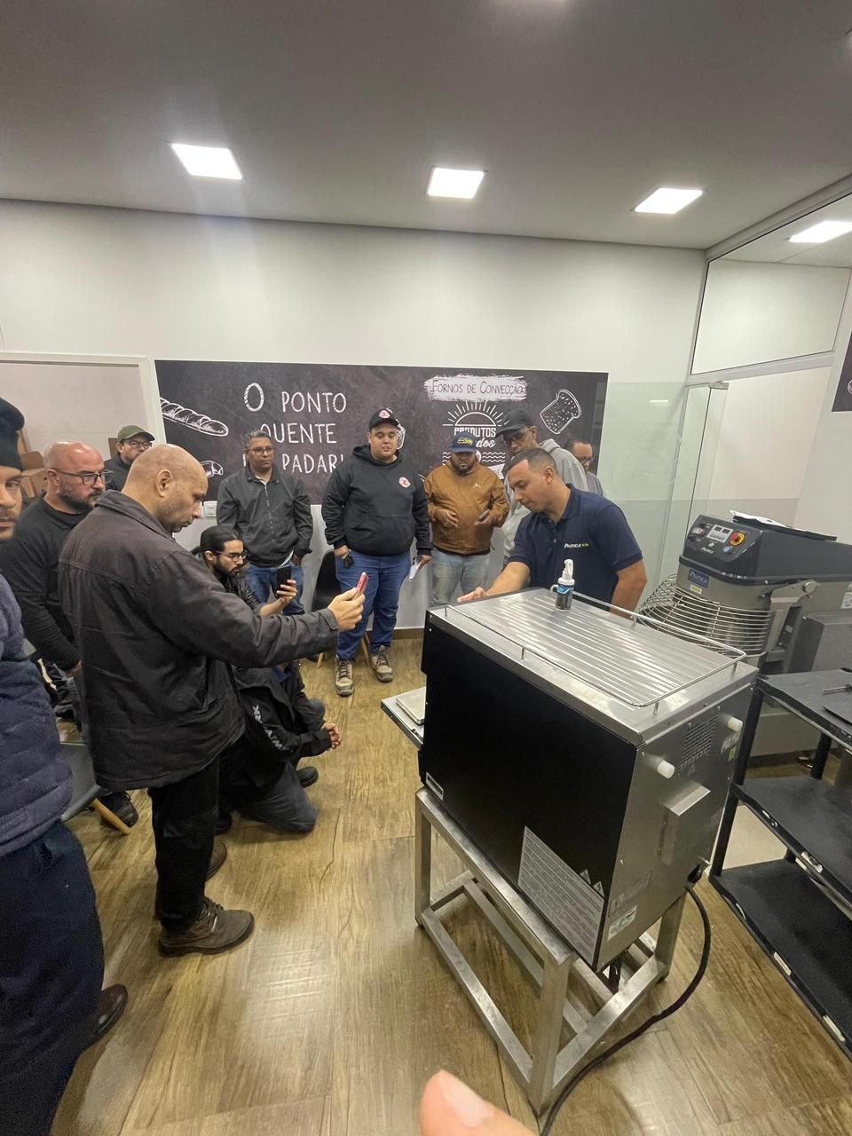 Grupo assiste à demonstração de uma máquina. Um homem aponta para ela. Ambiente de oficina com paredes brancas, iluminação e um quadro-negro.