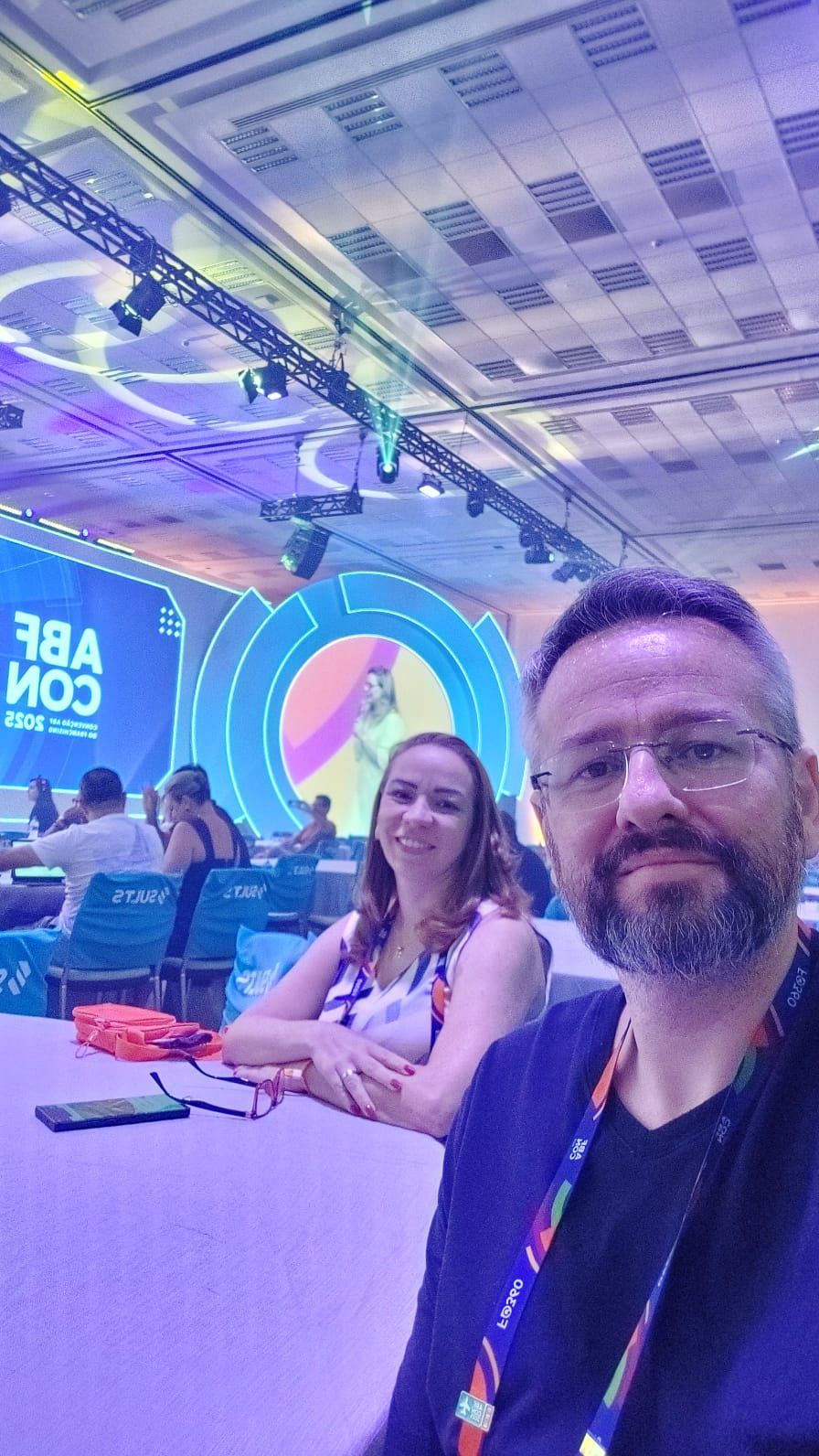 Duas pessoas em uma conferência. Homem de óculos, mulher com blusa florida, sentados à mesa. Fundo do palco visível.
