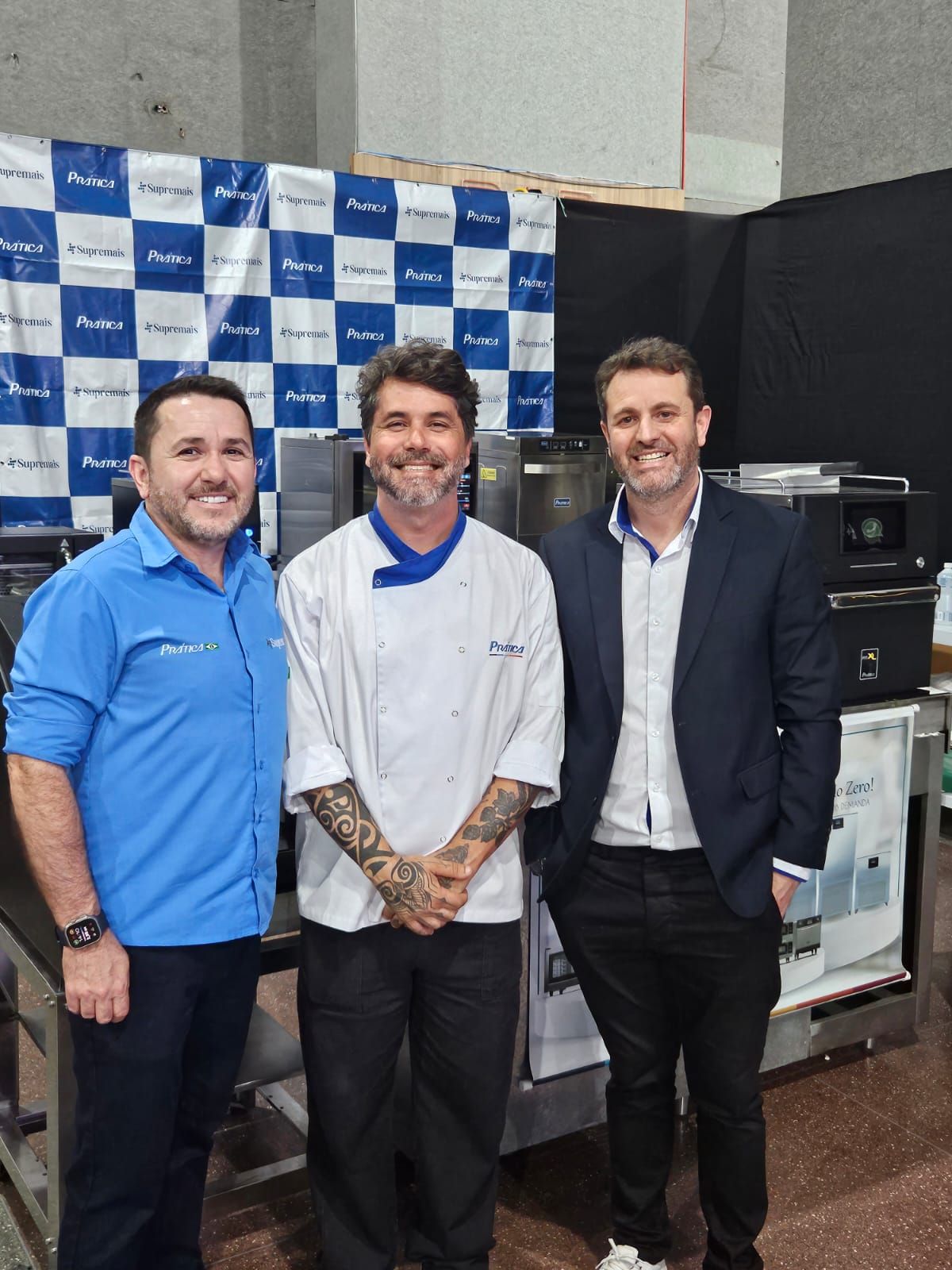Três homens em pé perto de equipamentos de cozinha, um vestindo uniforme de chef e dois em trajes casuais, sorrindo.