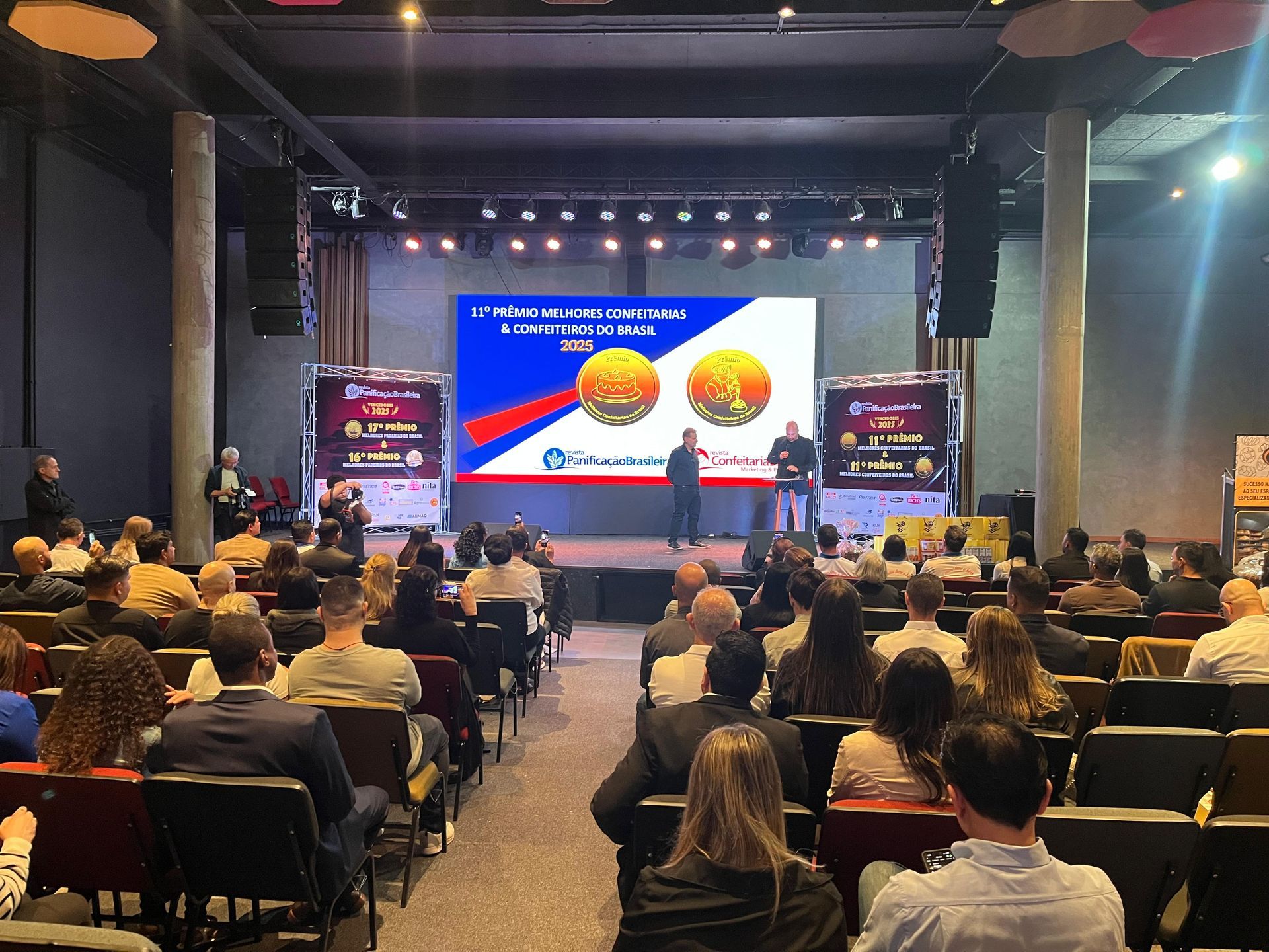 Público sentado em uma conferência; apresentadores em um palco com uma tela e um logotipo.