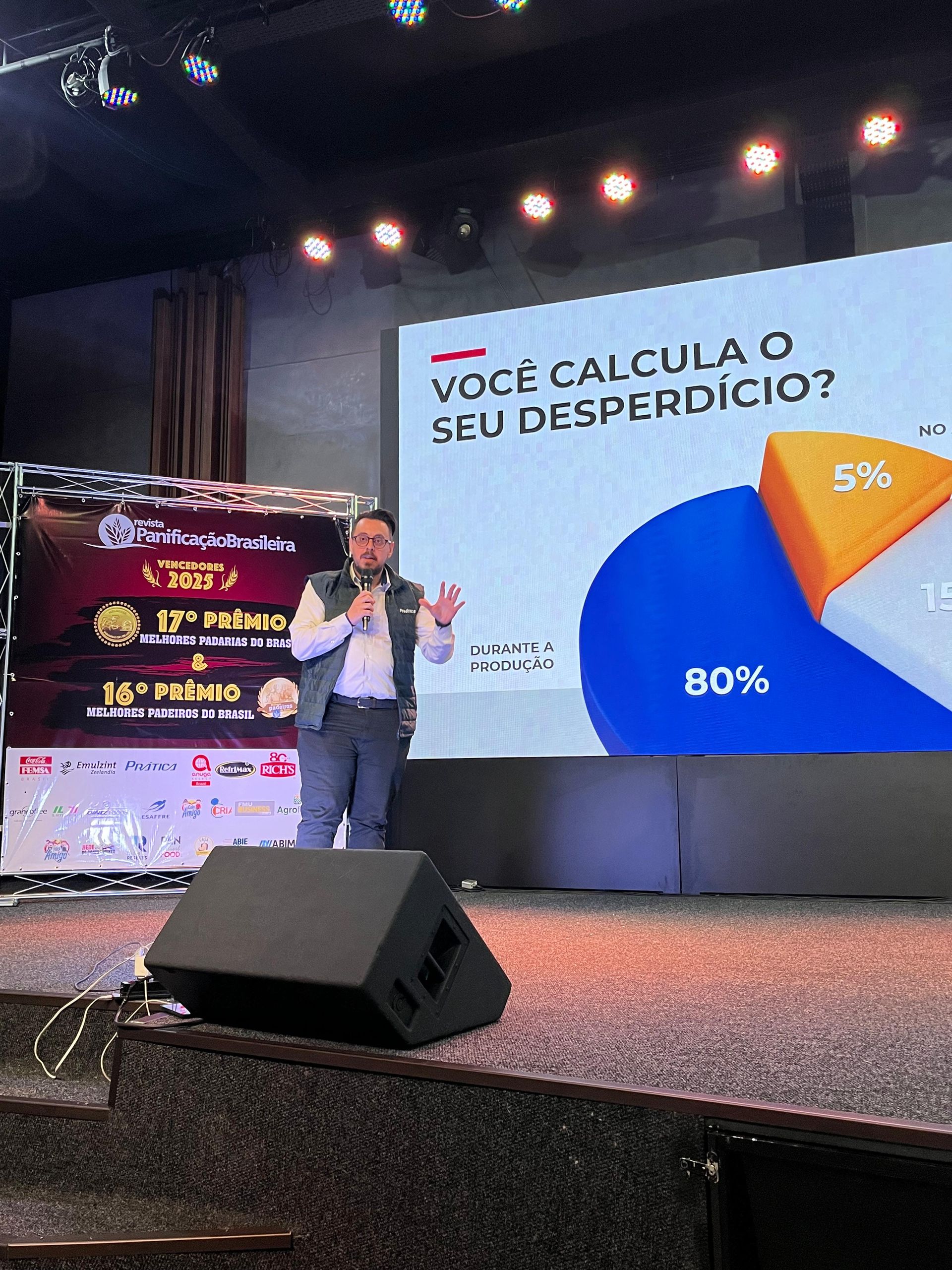 Homem no palco apresentando dados em uma tela, ao lado de banners do evento. Ele está gesticulando com as mãos.