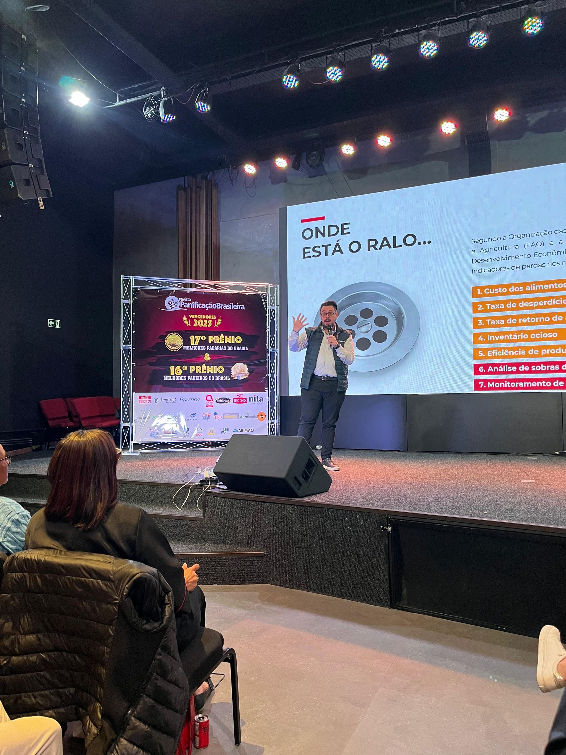 Homem apresentando no palco, telão com imagem, plateia em primeiro plano.