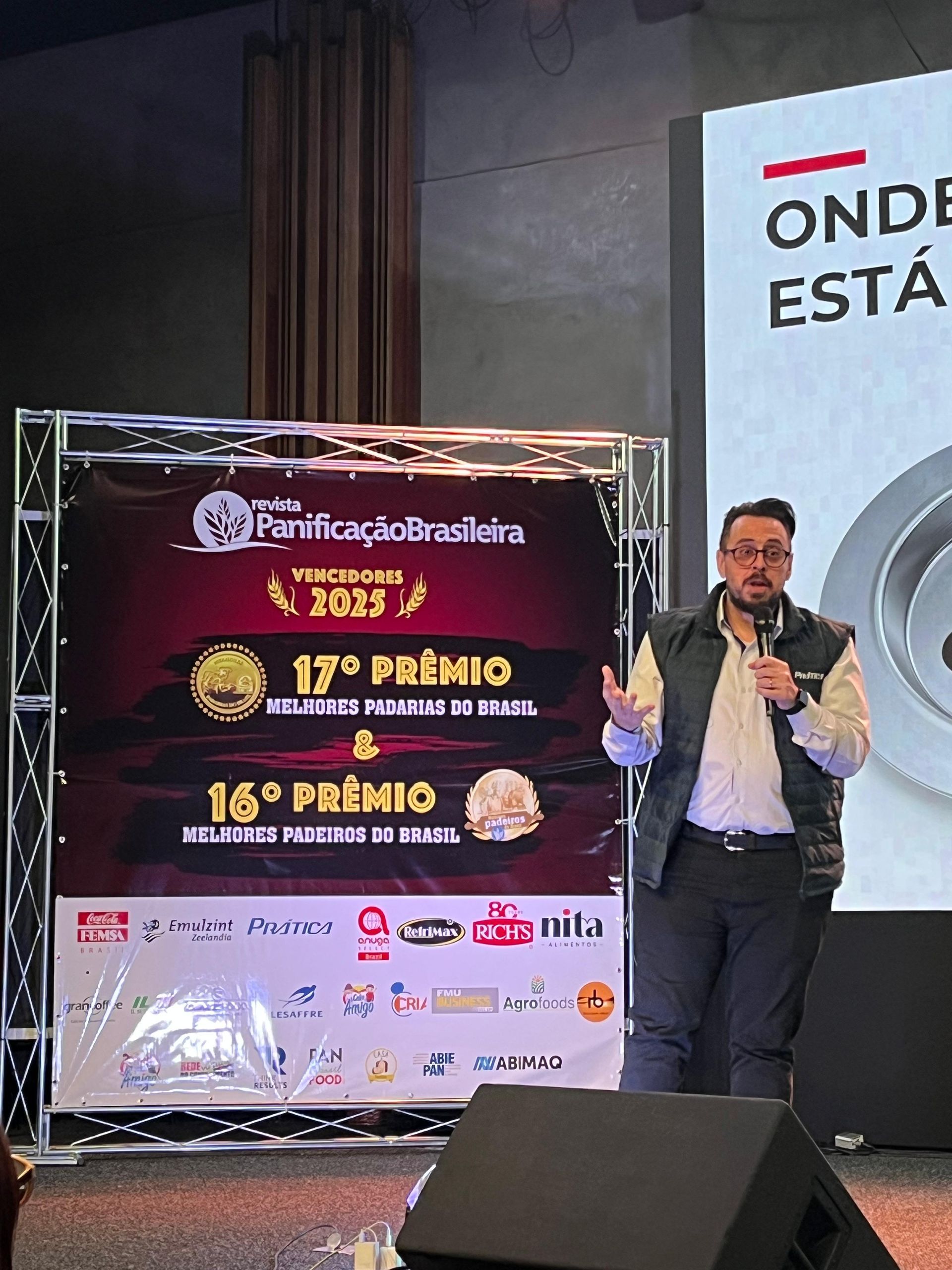 Homem discursando em um evento com um banner anunciando um prêmio. Palco montado em uma sala com uma tela de projeção.