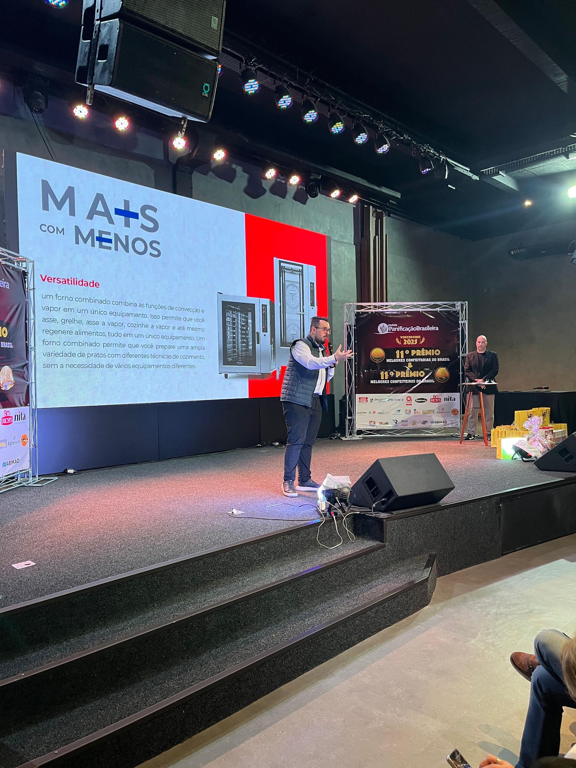 Homem no palco apresentando, com um telão atrás dele. O palco está iluminado, com uma placa escrito