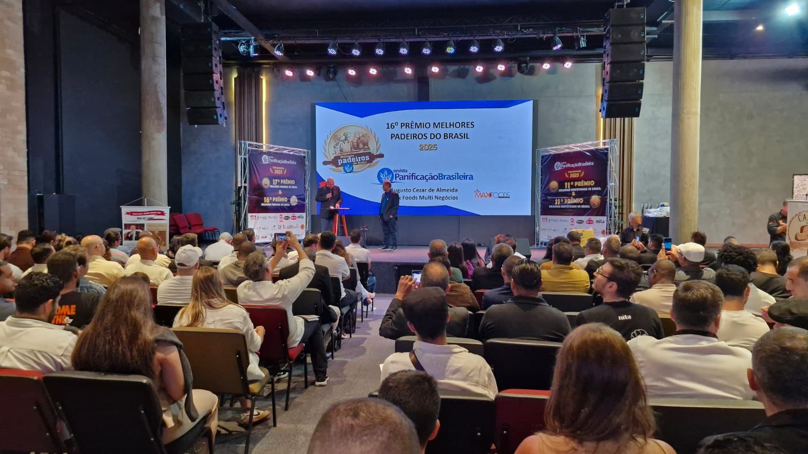 Um homem discursando em uma conferência. Tela grande com texto e uma plateia sentada em frente.