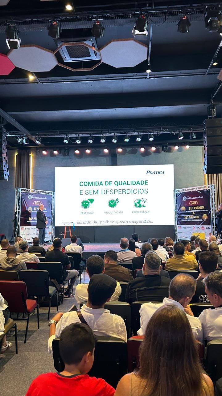 Conferência com apresentador no palco e plateia sentada. Um telão exibe texto e gráficos.