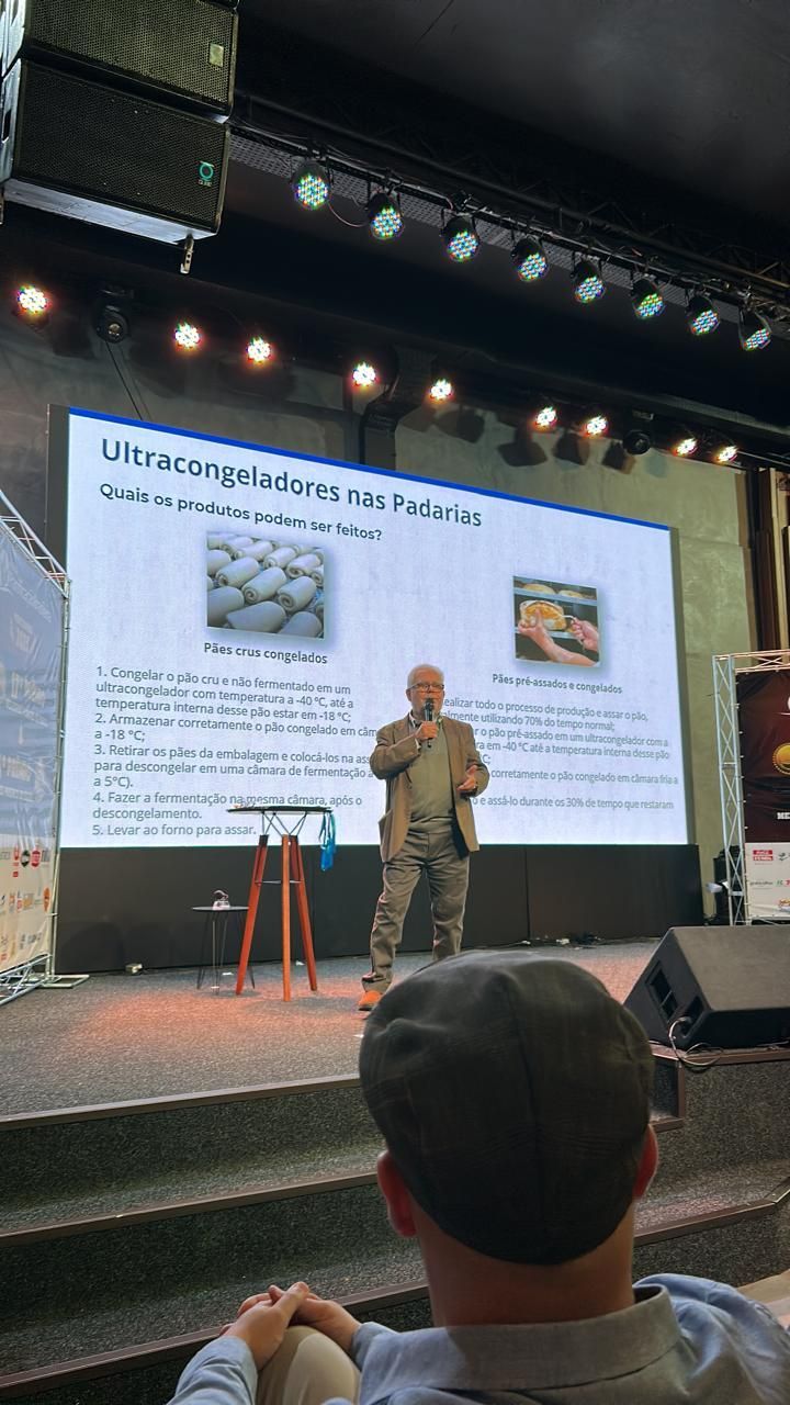 Homem apresentando em um palco em frente a uma tela com texto e imagens. Membro da plateia em primeiro plano.