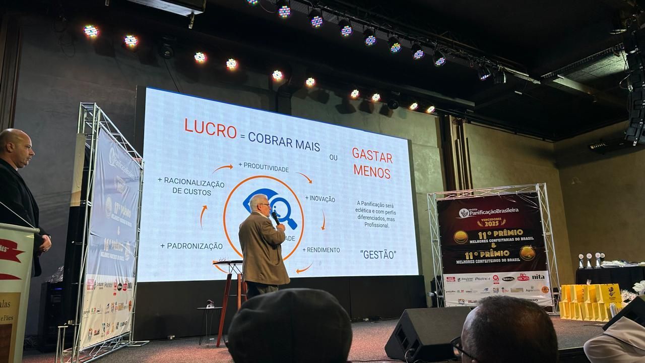 Homem discursando no palco, apresentação exibida em um telão. Banner do evento, público sentado.