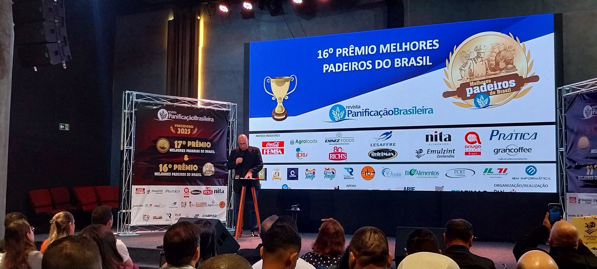 Uma pessoa discursa no palco durante um evento. Um telão exibe logotipos e texto. O público está sentado.