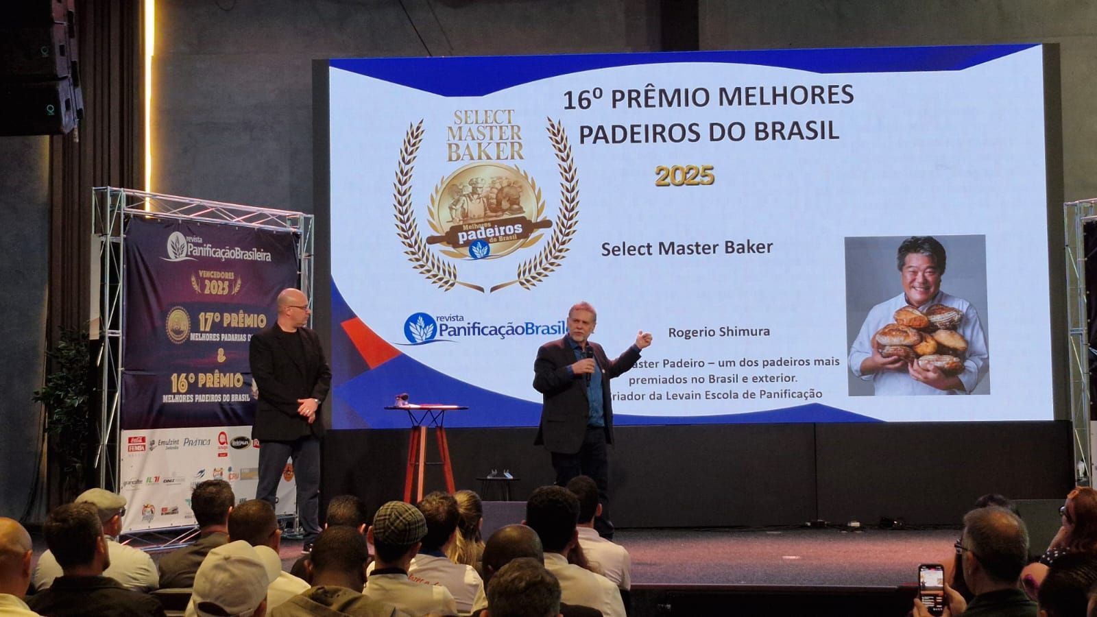 Dois homens no palco durante uma cerimônia de premiação de confeitaria. Um telão exibe o logotipo do evento e a foto de um confeiteiro. A plateia está em primeiro plano.