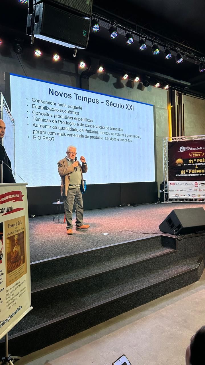 Homem discursando em um palco com um telão exibindo texto. O palco possui degraus, uma faixa e caixas de som.