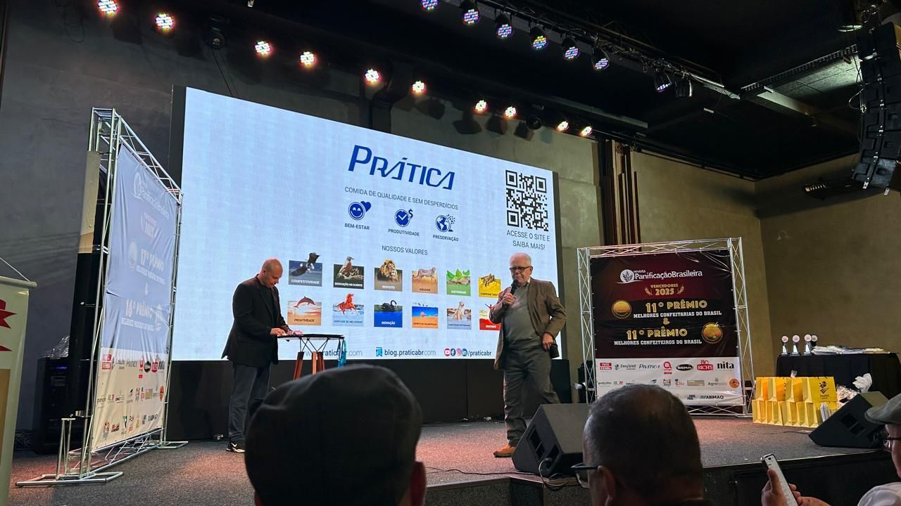 Dois homens no palco apresentando em frente a uma tela com um logotipo e elementos gráficos. Cenário de evento.