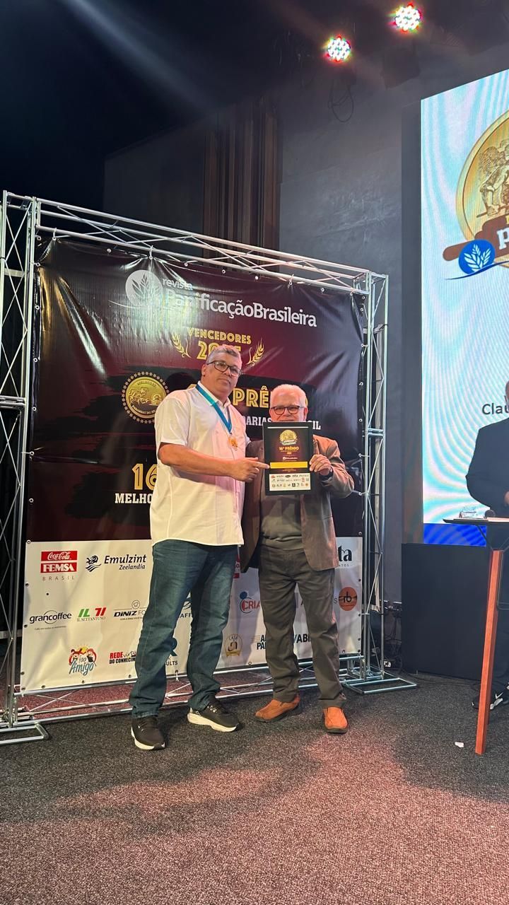 Dois homens em um evento, um deles recebendo um prêmio. Ao fundo, um telão e banners.