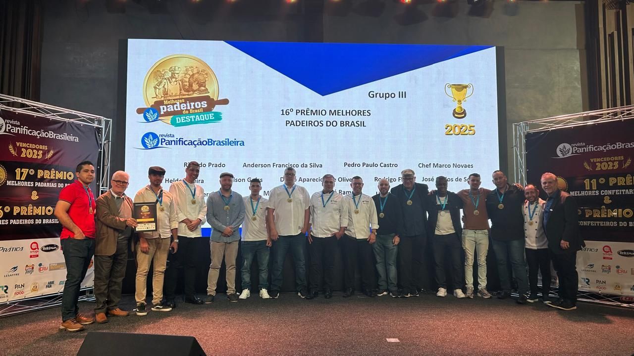 Grupo de pessoas posando em um palco, com um telão exibindo logotipos e texto, provavelmente uma cerimônia de premiação.
