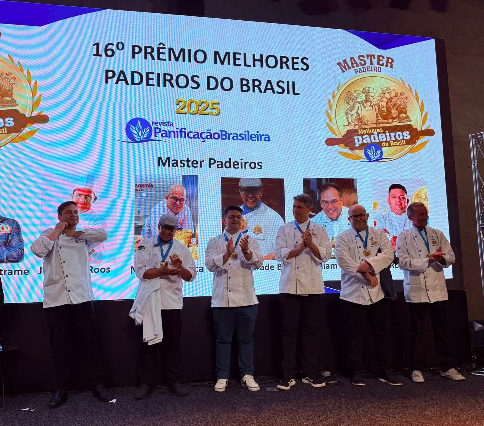 Grupo de padeiros de jaleco branco no palco, aplaudindo durante uma cerimônia de premiação. Um telão exibe o título e o logotipo do evento.