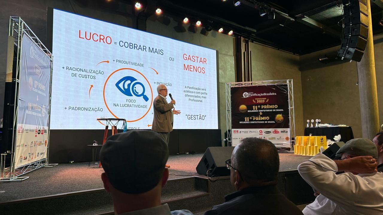 Um homem dá uma palestra em uma conferência, gesticulando em direção a uma tela com texto e gráficos; os membros da plateia estão sentados.