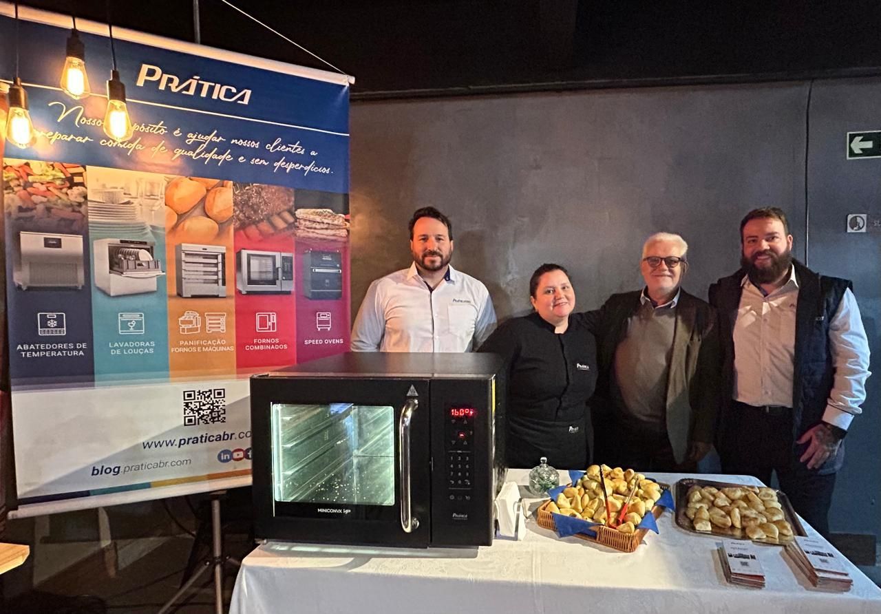 Pessoas em uma apresentação de comida com um forno Proma e banner. Mesa de exposição com alimentos e equipamentos.