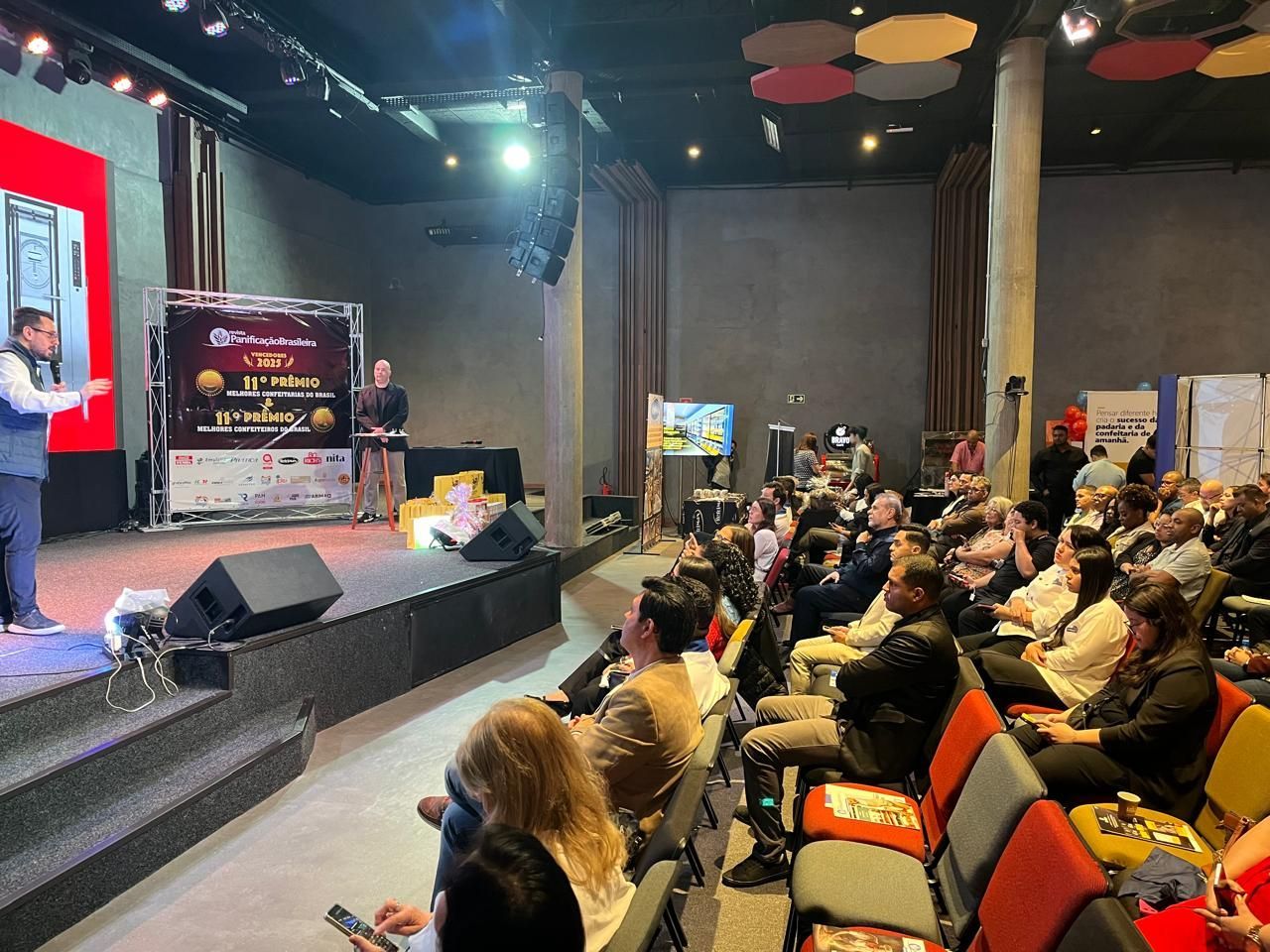 Homem discursando no palco durante evento, plateia sentada, banner ao fundo, sala ampla.