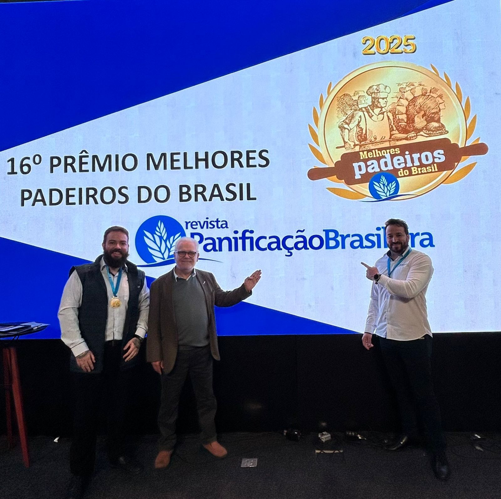 Três homens em uma cerimônia de premiação, em pé diante de uma tela com texto e um logotipo. Um deles aponta para a tela.