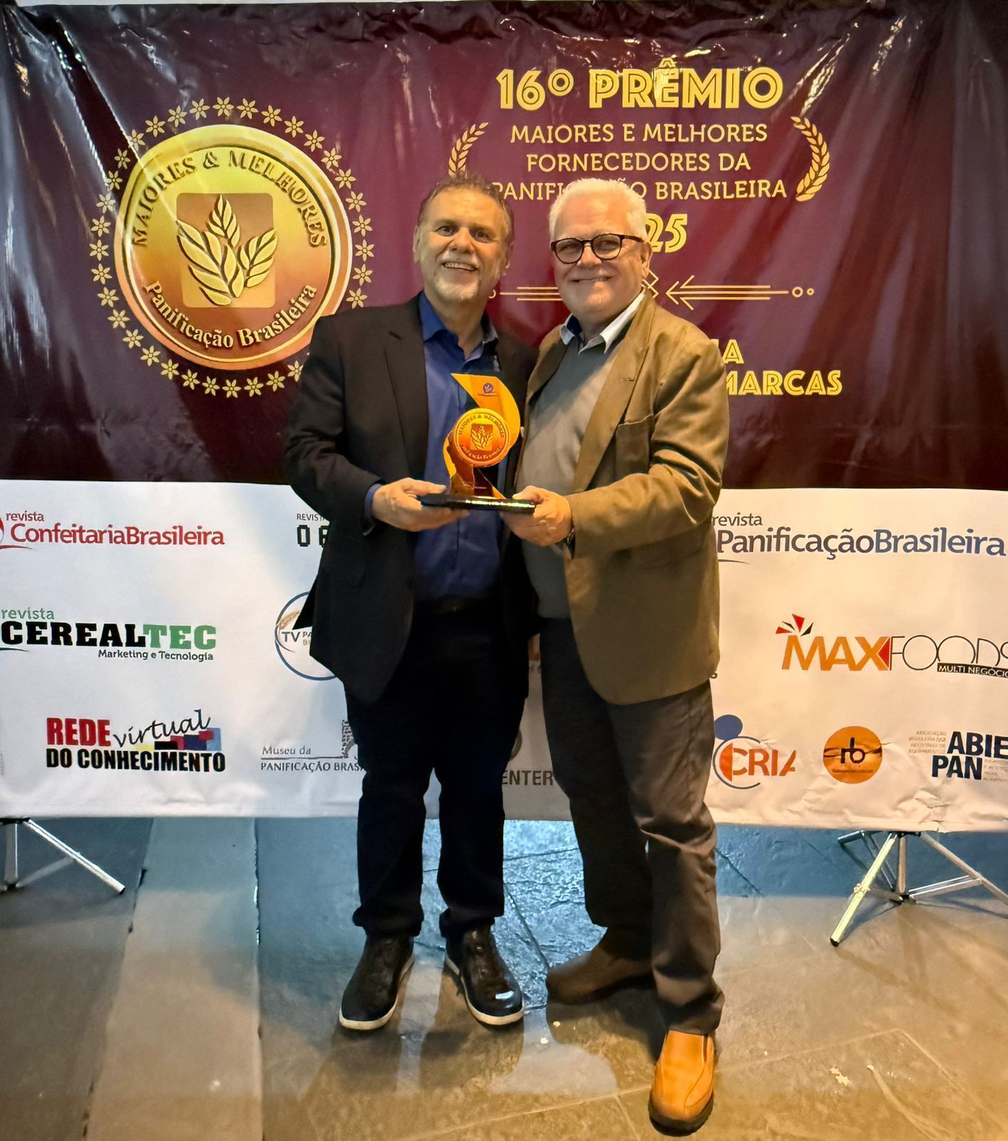 Dois homens segurando uma condecoração; um de blazer, o outro de jaqueta, em frente a uma faixa.