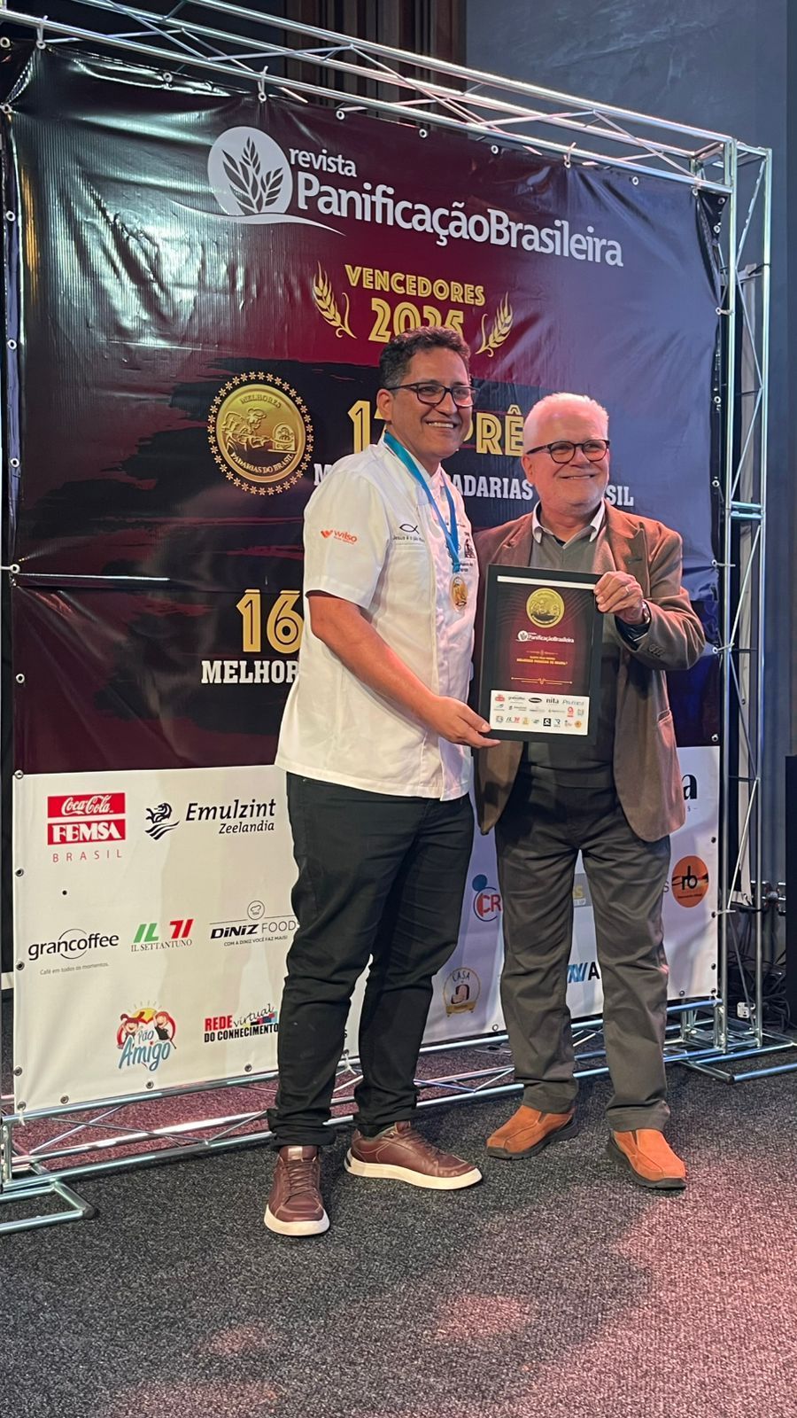 Dois homens segurando um prêmio e posando, sorrindo, em frente a uma faixa durante um evento.