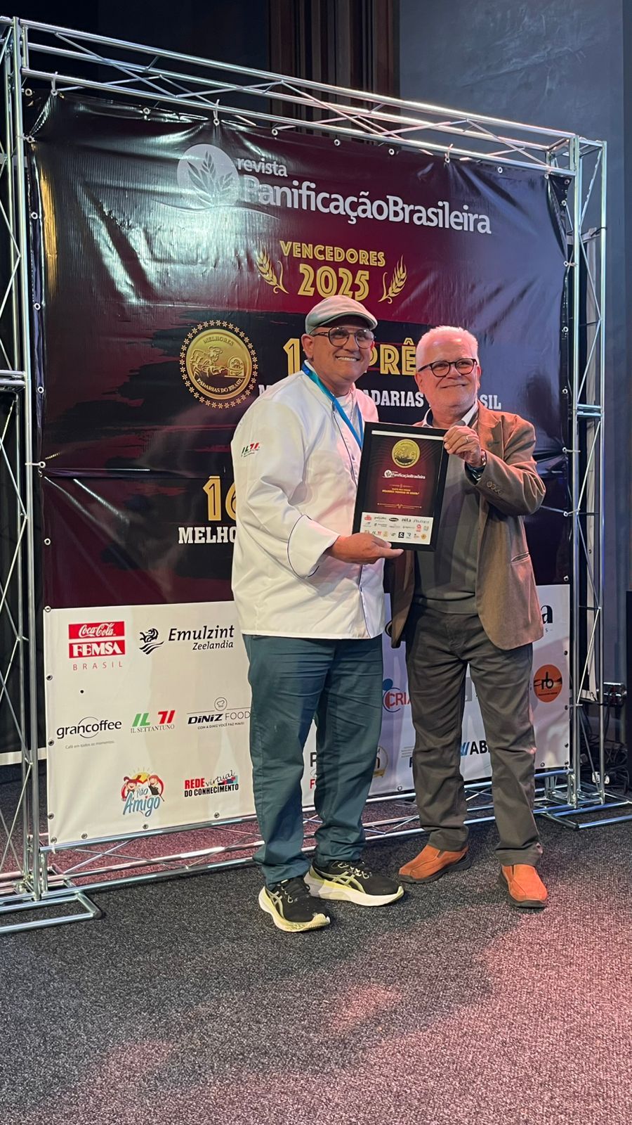 Chef de cozinha, vestindo jaleco branco e calça jeans, recebe prêmio de um homem de blazer. O cenário da cerimônia é visível ao fundo.