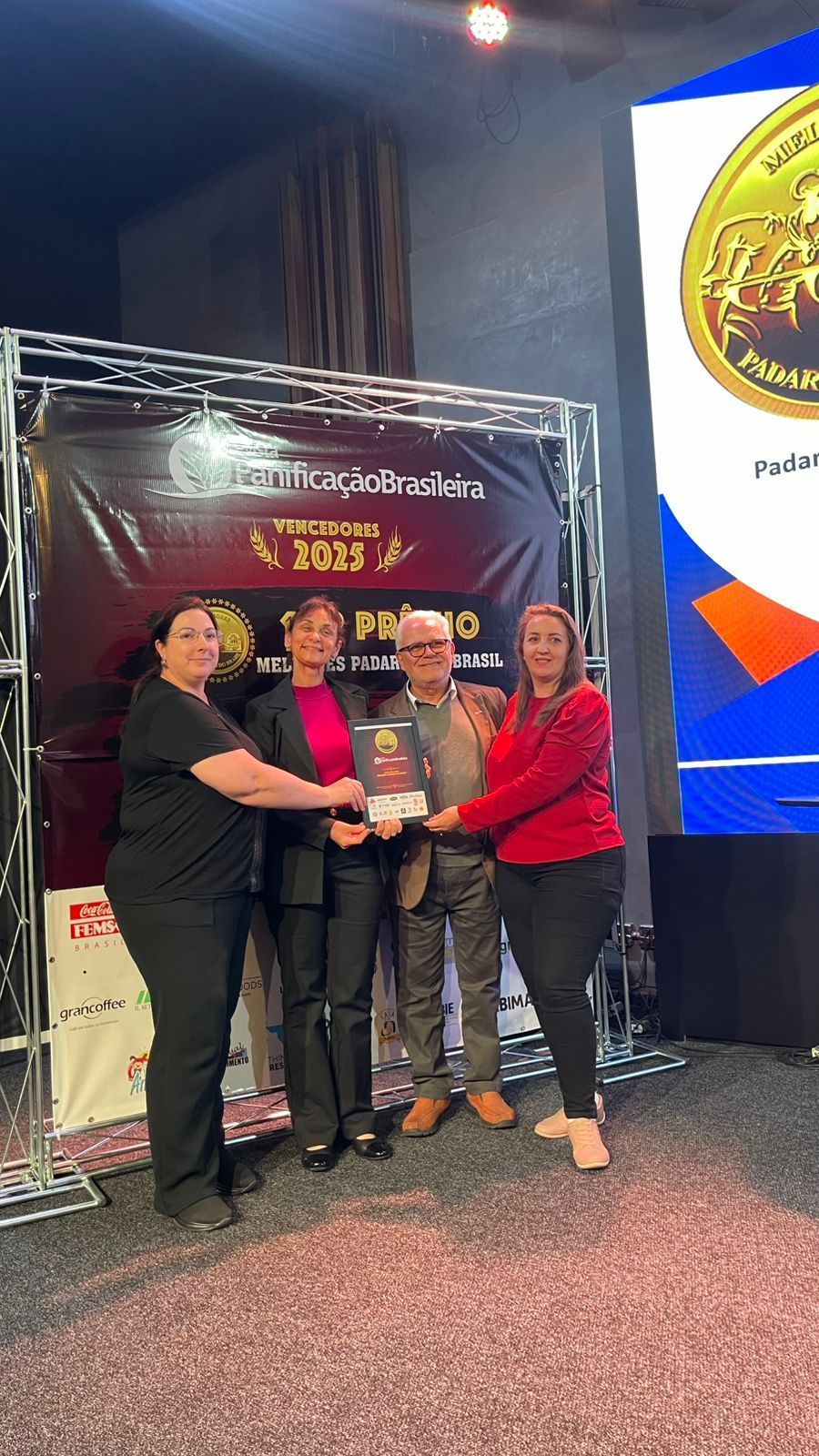 Quatro pessoas segurando um certificado em uma cerimônia de premiação, em pé em frente a uma faixa.