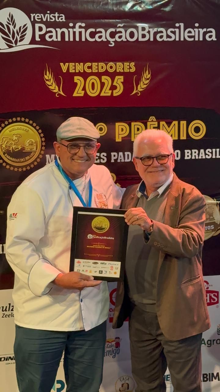 Homem de uniforme de chef segurando um prêmio emoldurado ao lado de um homem de blazer; cenário de evento de premiação com faixa.
