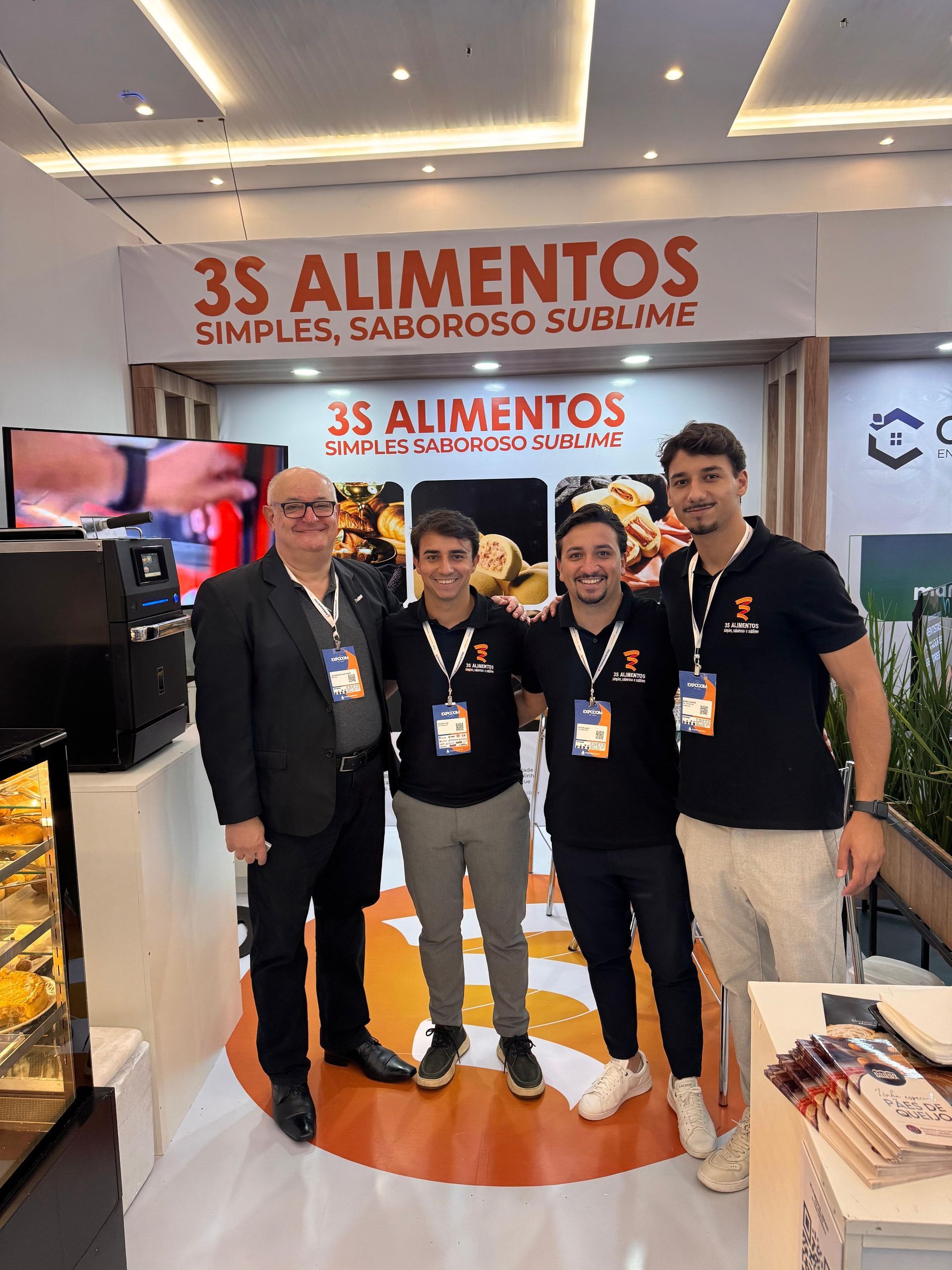 Quatro pessoas posam em um estande da 3S Alimentos com um logotipo que apresenta o nome da marca.