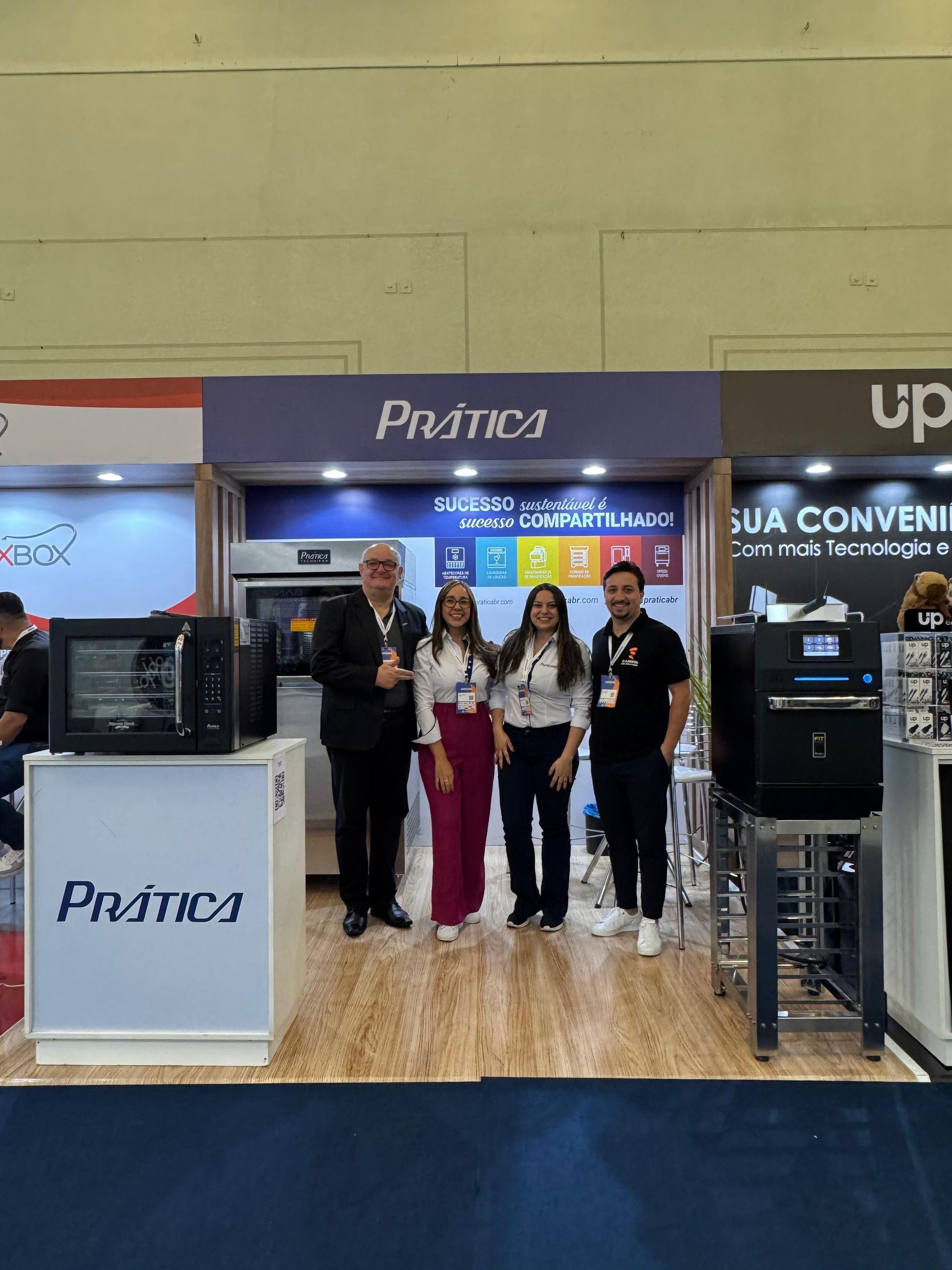 Pessoas posam para fotos em frente a um estande da Prática com fornos em uma feira comercial.