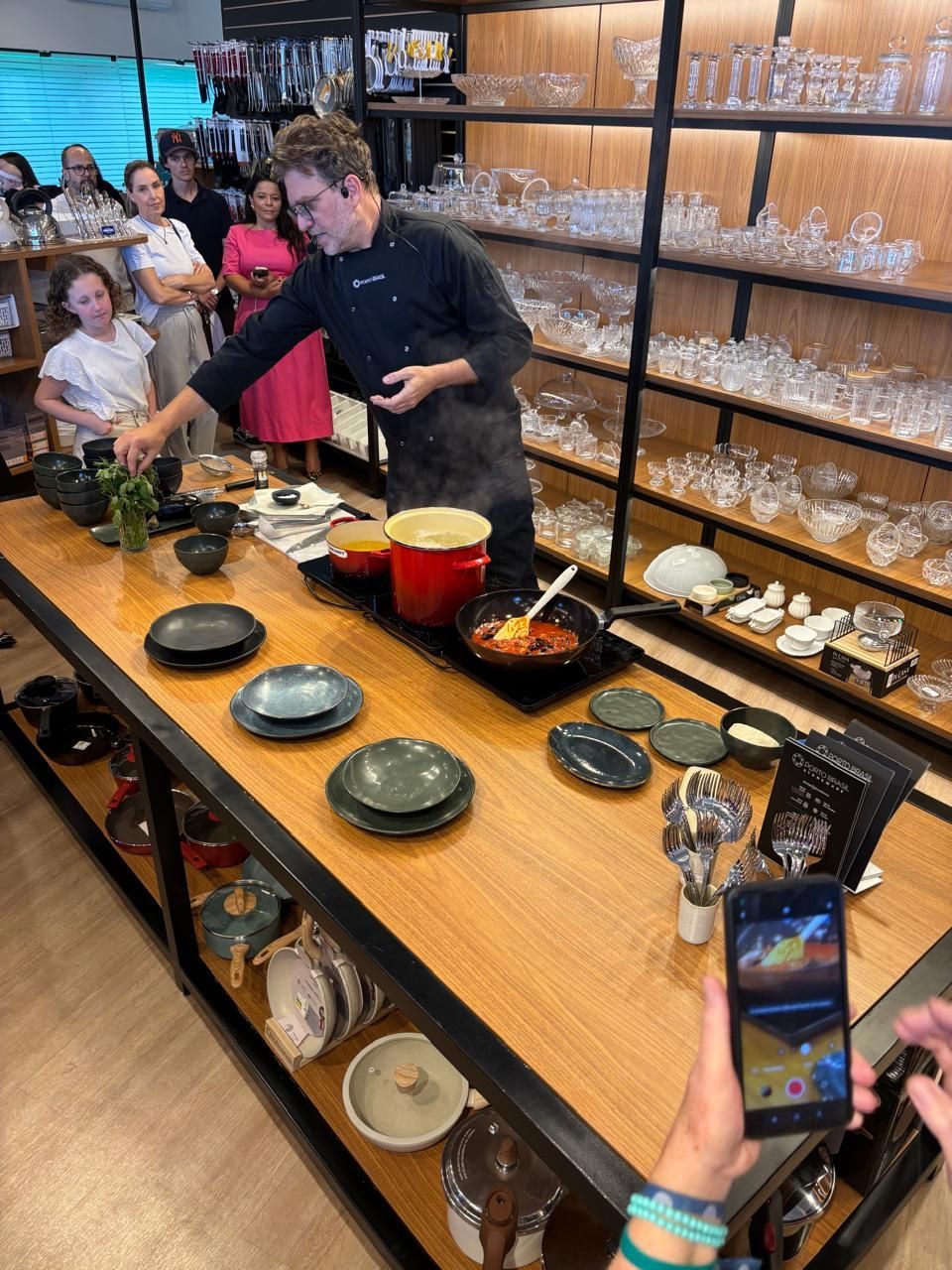 Chef demonstra o preparo de uma receita em uma mesa, utilizando utensílios de cozinha, enquanto o público observa em uma loja de varejo.