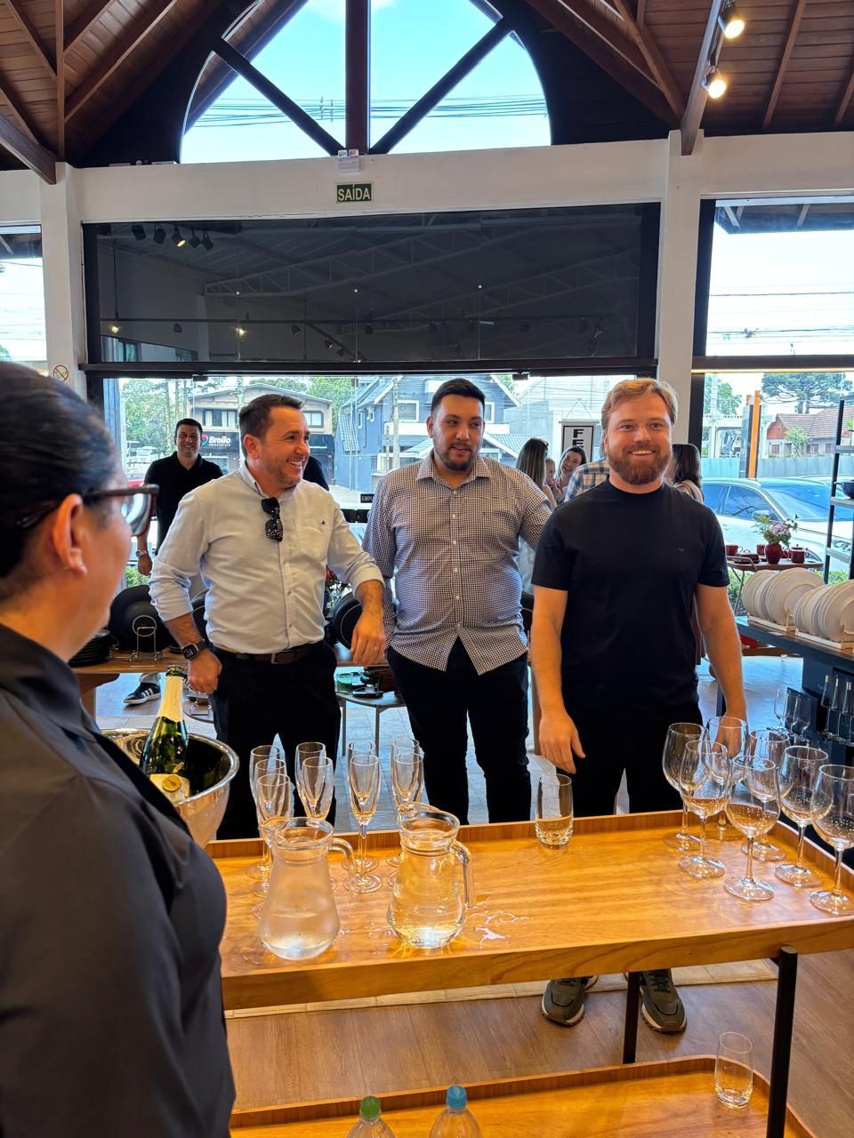 Três homens estão em pé junto a um balcão de madeira com copos e taças; uma mulher está de frente para eles. Interior com grandes janelas.