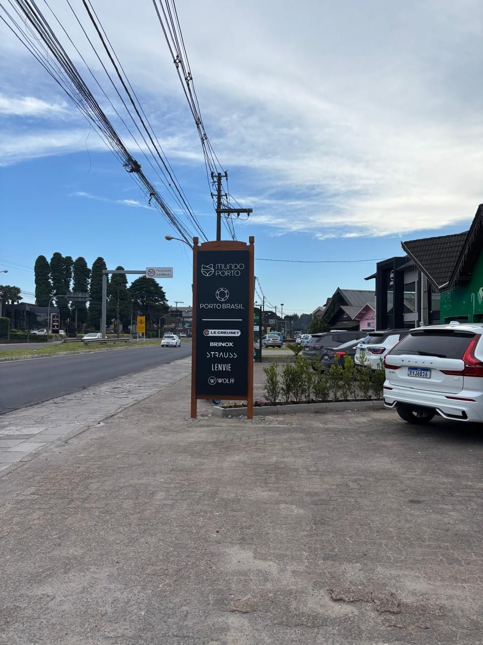 Placa com o nome da empresa à beira da estrada, no asfalto, na via, em carros, postes de luz, árvores e céu azul.