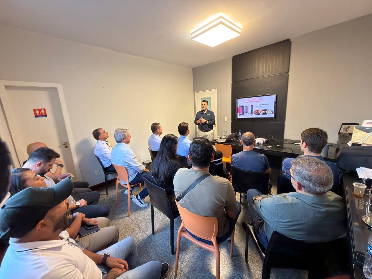 Um homem fazendo uma apresentação para um grupo em uma sala com uma tela. As pessoas estão sentadas e assistindo.