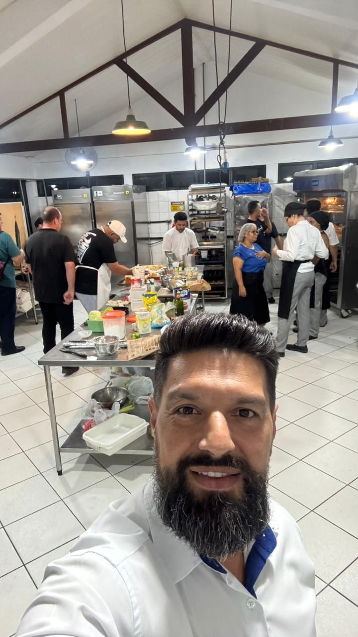 Homem de camisa branca sorri enquanto tira uma selfie em uma cozinha grande com funcionários cozinhando.