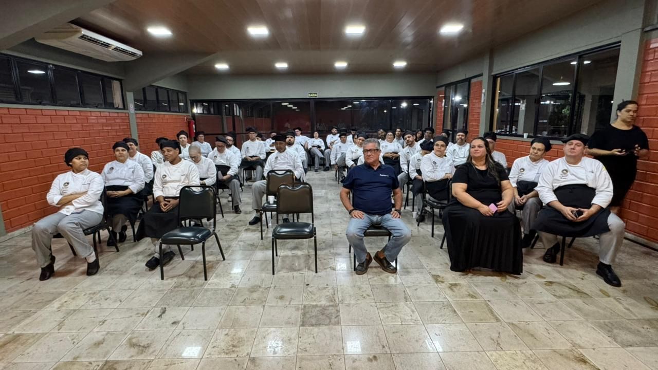 Pessoas vestindo jalecos brancos e calças pretas, sentadas em fileiras, escutavam em uma sala com paredes de tijolos.