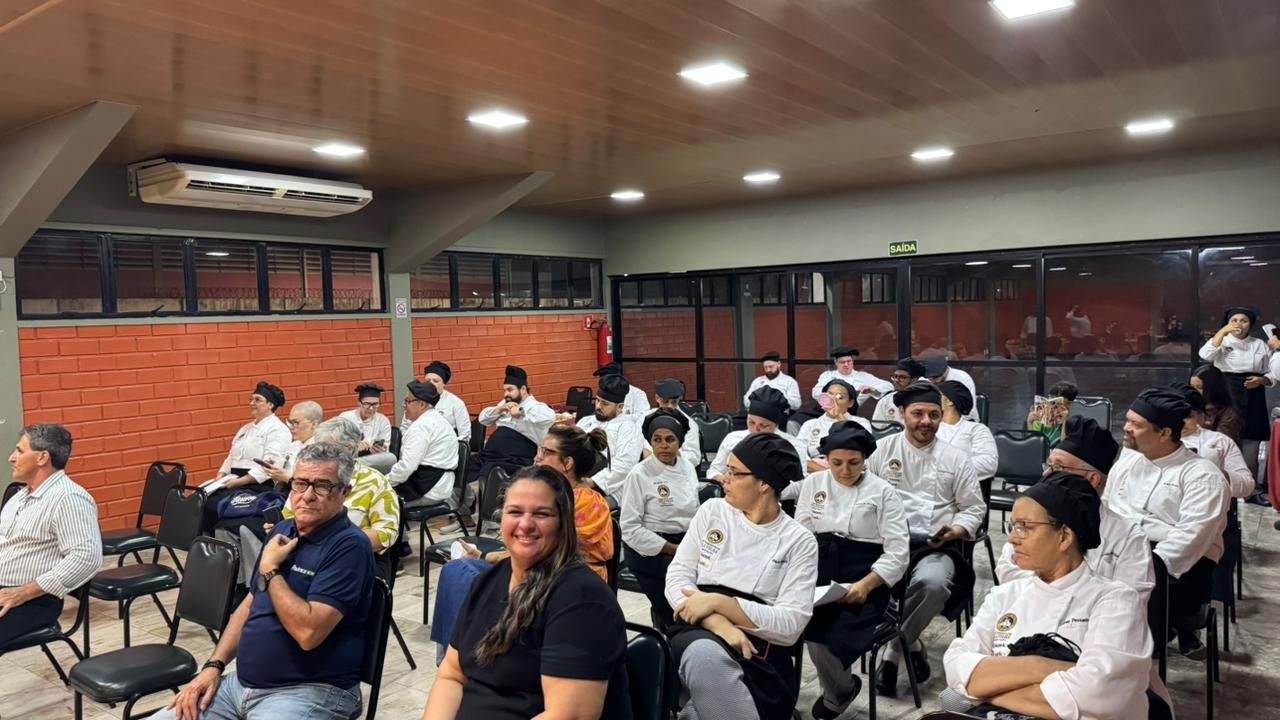Pessoas sentadas em uma sala, algumas vestindo uniformes de chef, participando de uma reunião. 