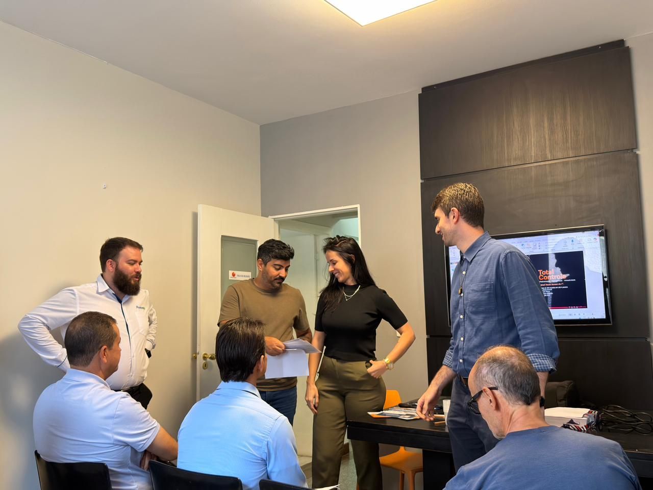 Pessoas reunidas em um escritório. Algumas estão de pé, outras sentadas. Uma mulher segura papéis e gesticula.