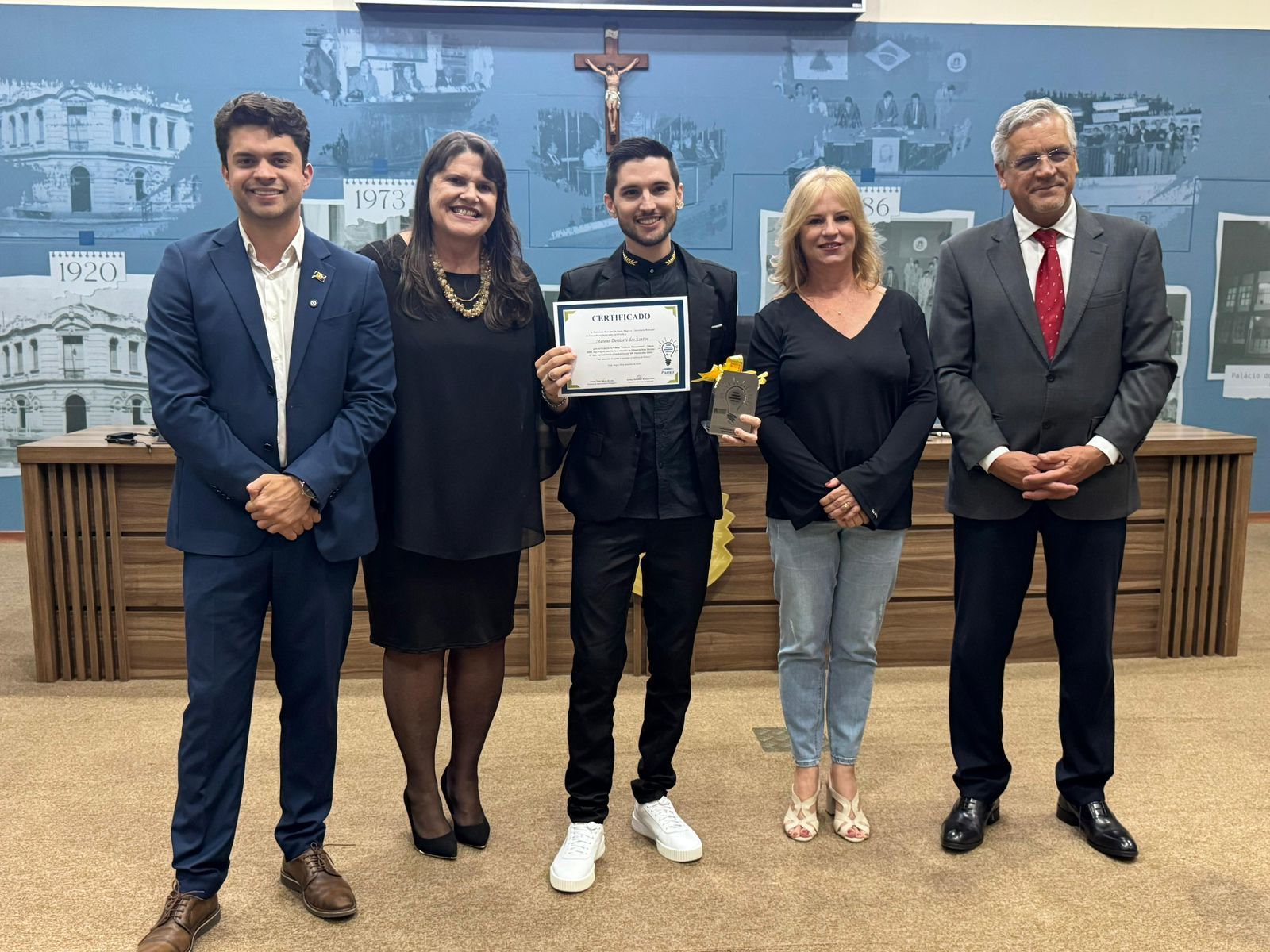 Grupo de cinco pessoas em um evento; um homem segura um certificado e um pequeno troféu. Eles estão em frente a um painel de madeira com imagens.