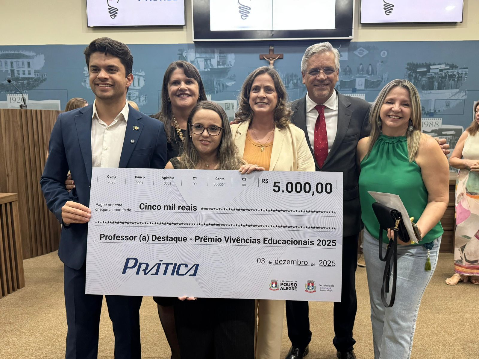 Um grupo segura um cheque simbólico de 5.000,00. As pessoas estão em uma sala, comemorando o prêmio.