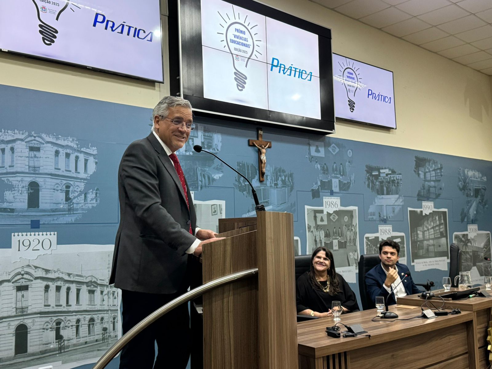Homem discursando em um pódio com duas pessoas sentadas, em frente a uma tela com o logotipo de uma lâmpada.