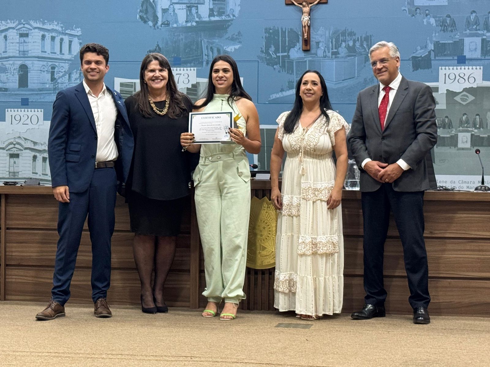 Cinco pessoas estão de pé, segurando um certificado. Elas estão em uma sala com uma cruz e prédios na parede.