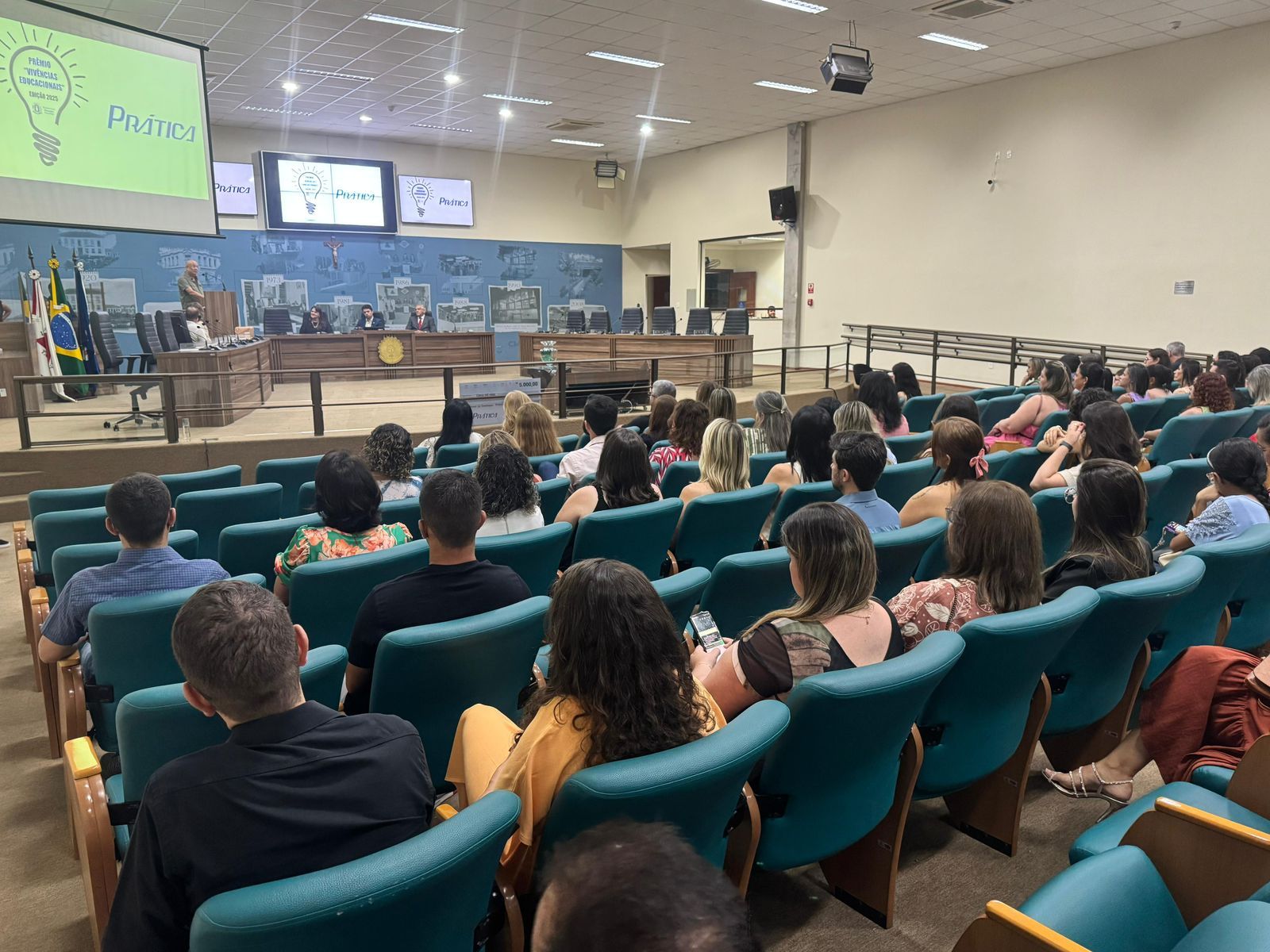 Público em um auditório, de frente para um palco com uma tela exibindo texto e um logotipo, presumivelmente assistindo a um evento.