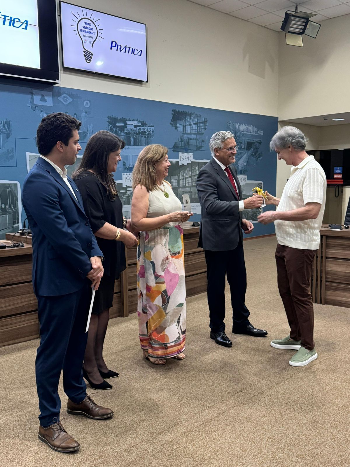 Grupo de pessoas em um ambiente de escritório. Um homem está recebendo um prêmio, outros observam e um deles segura um microfone.