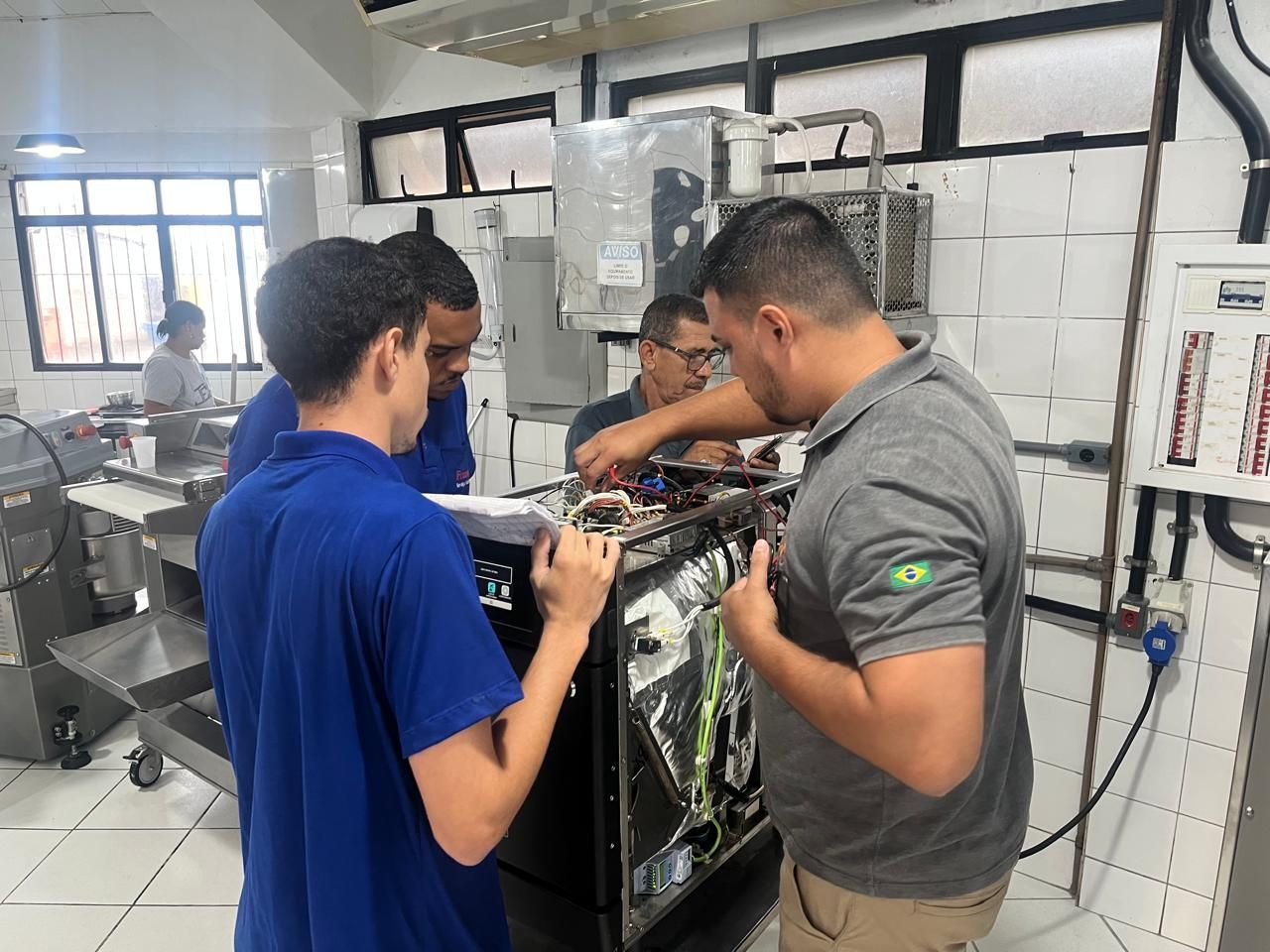 Quatro homens em um laboratório examinam componentes elétricos de uma máquina. Um deles lê um documento.