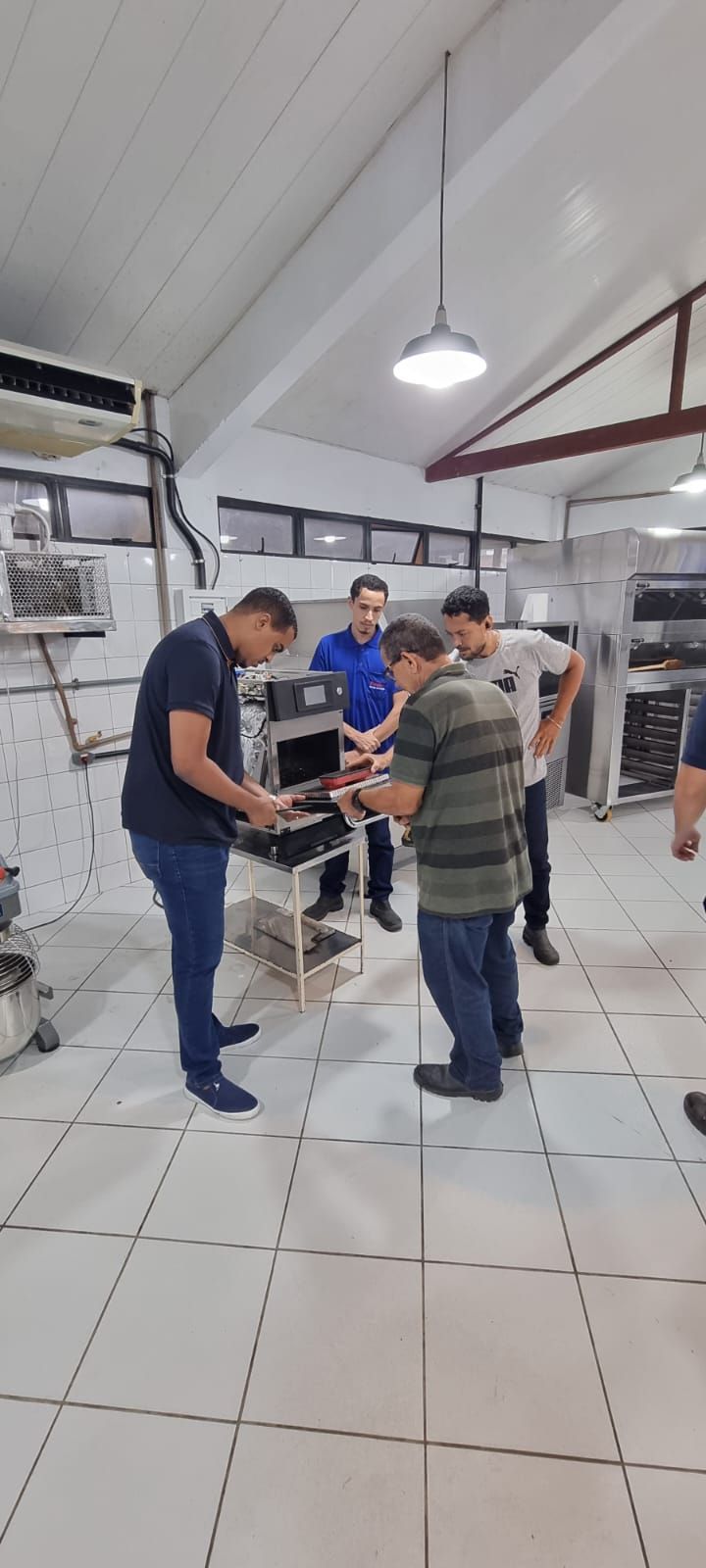 Quatro homens examinam equipamentos em uma sala com azulejos brancos e elementos industriais; iluminação no teto.