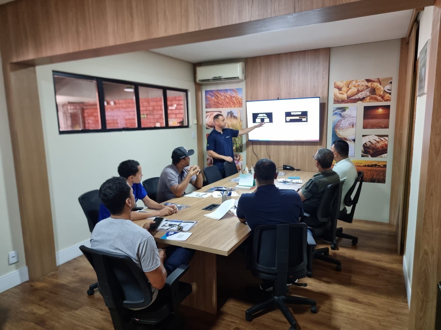 Homem fazendo apresentação em reunião para um grupo sentado à mesa de conferência.